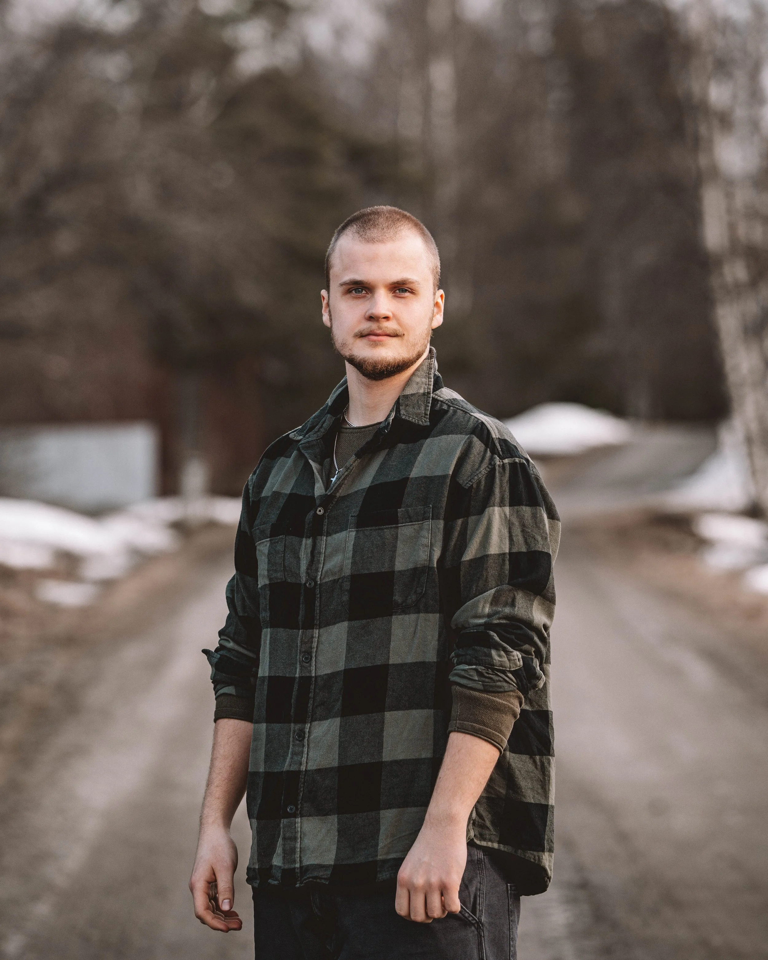 En ung man står på en grusväg i en skog i Örnsköldsvik, Höga Kusten, klädd i en svart och grön rutnätsflanellskjorta, med träd och snö i bakgrunden.