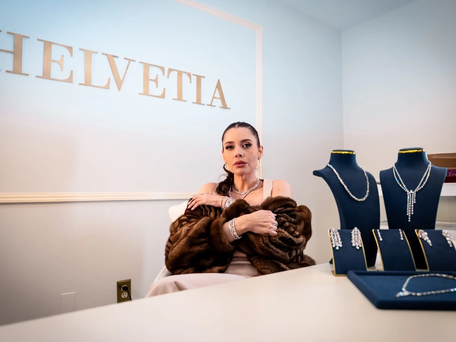 Glroia sitting at a jewelry display table, wearing a brown fur coat and jewelry, with jewelry on display on black busts and stands on the table, and a sign that reads 'HELVETIA' on the wall behind her.