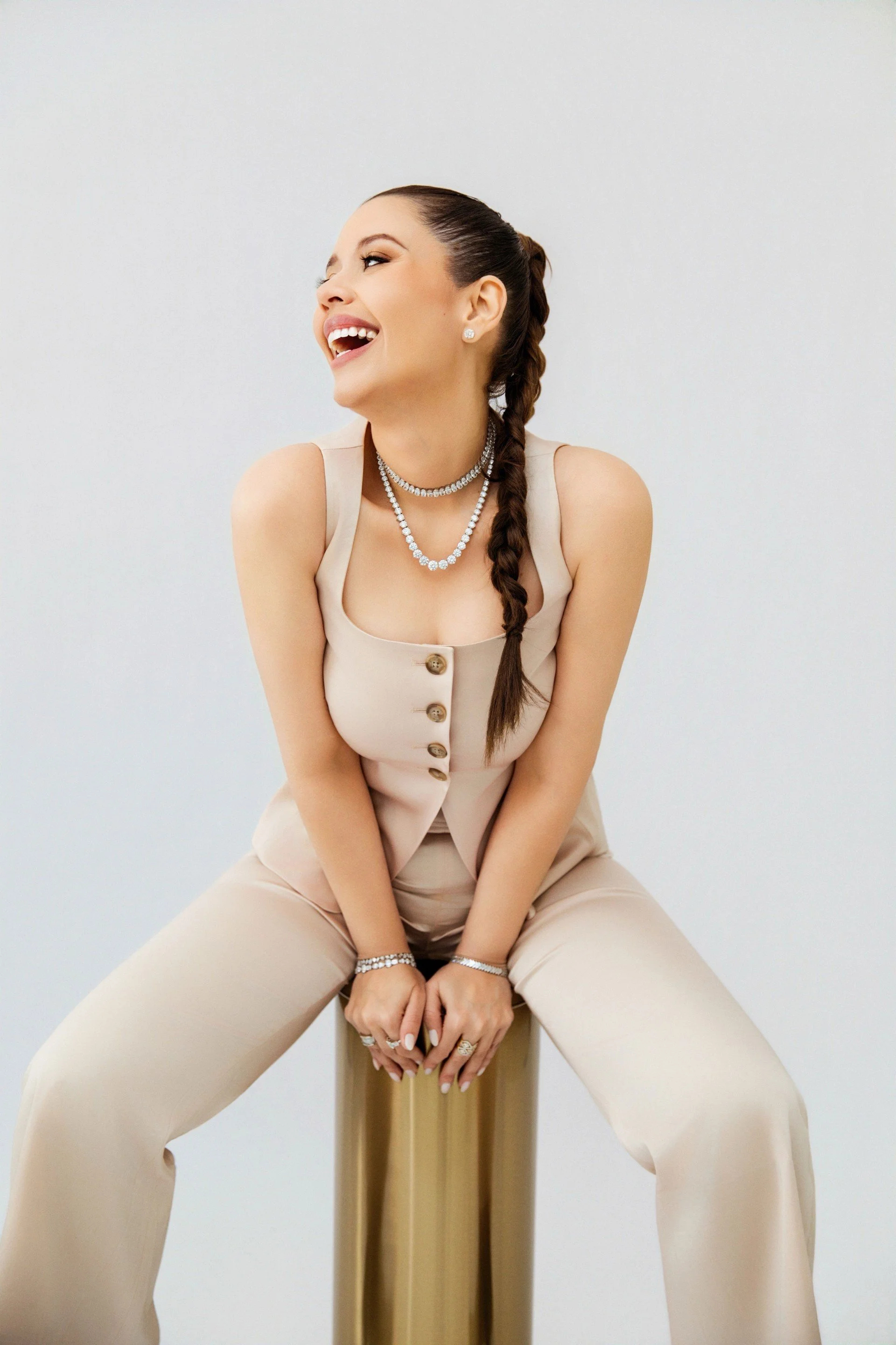A Gloria sitting on a gold-colored stool against a white background, wearing a beige sleeveless top and matching wide-leg pants, accessorized with layered pearl necklaces, earrings, and bracelets.