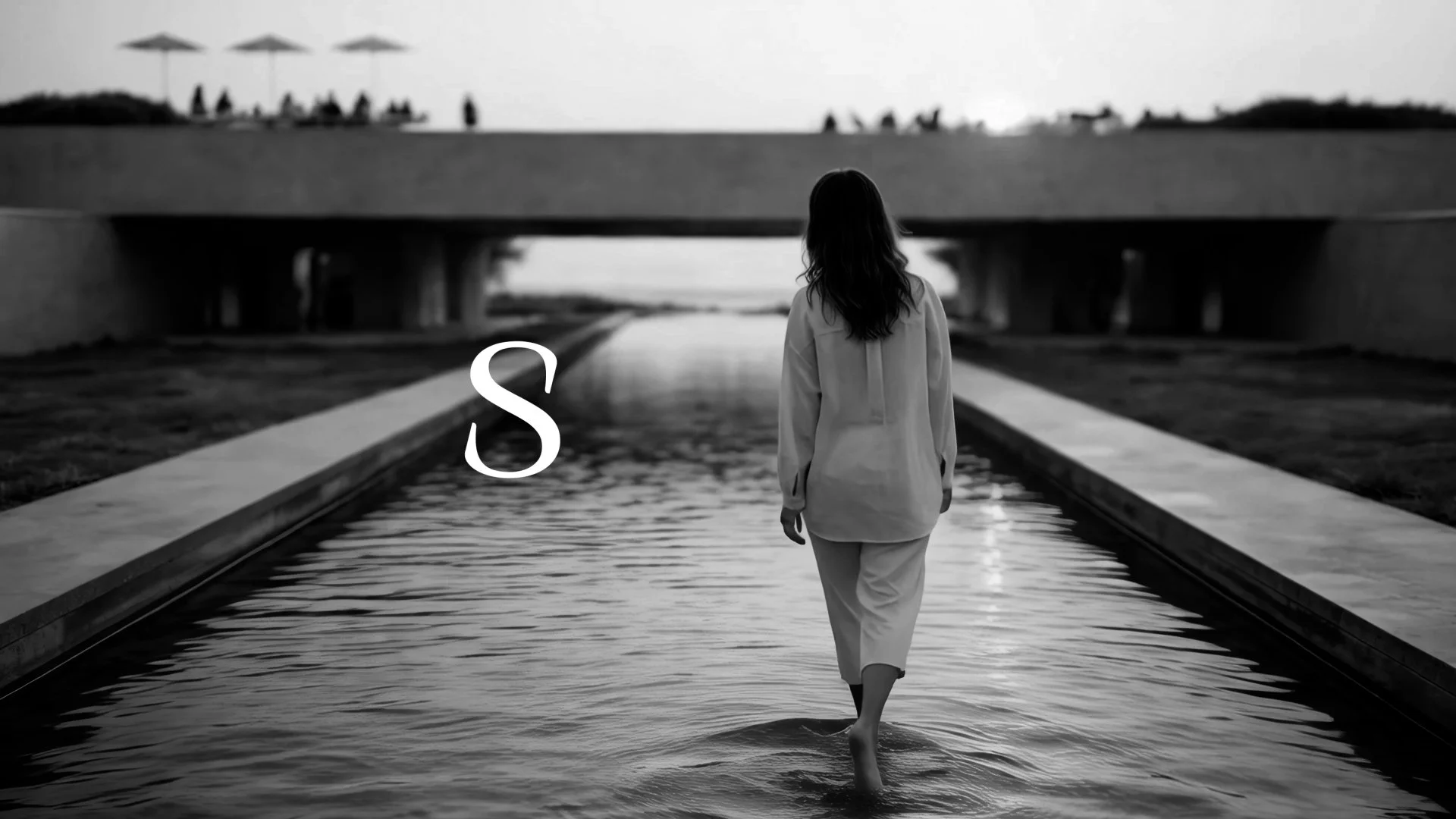 A woman with long hair walking through shallow water in an outdoor setting, with a bridge and people in the background, in black and white.