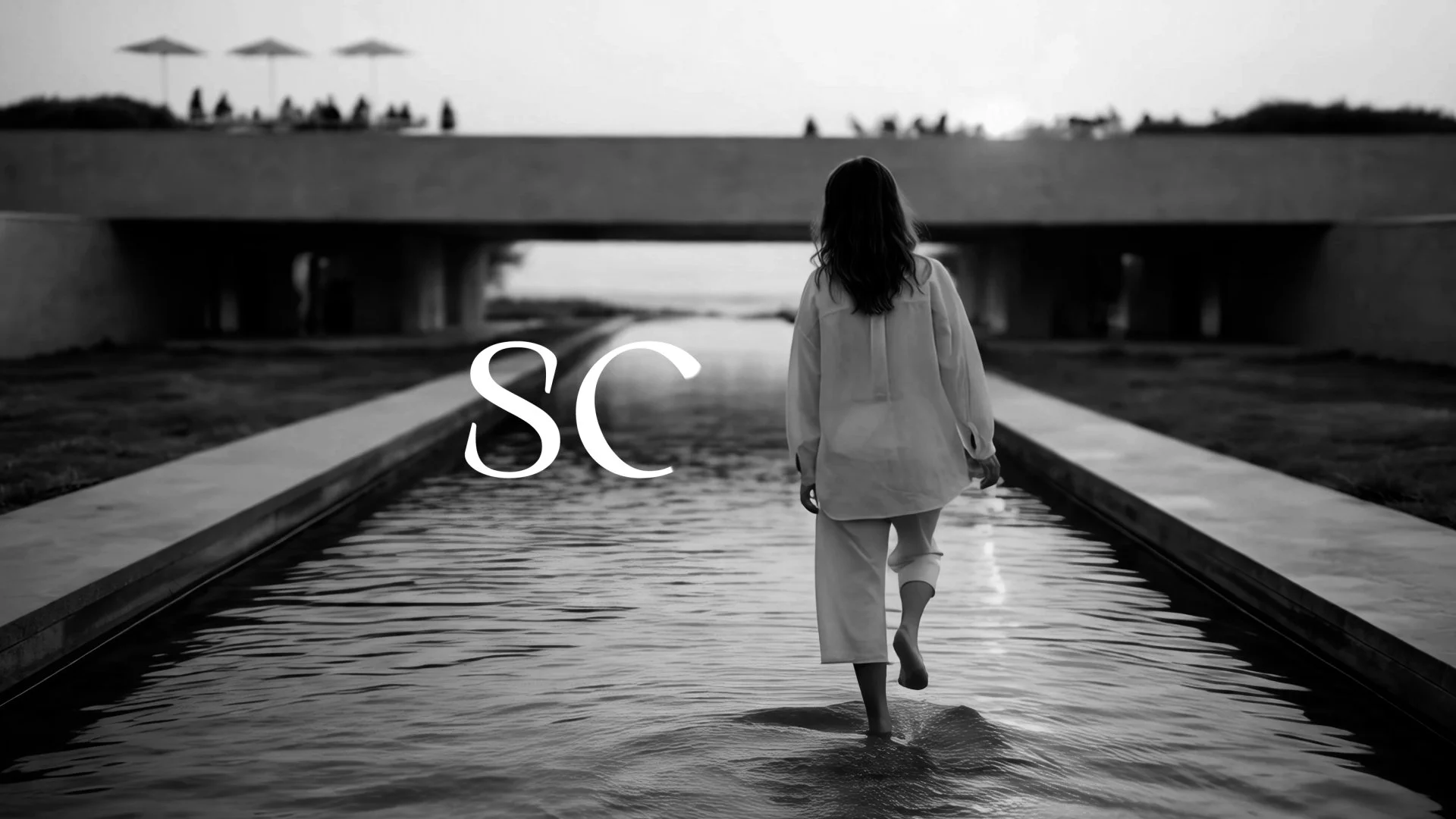 A woman with long hair walking barefoot through a shallow water feature on a pathway near a bridge, with trees and umbrellas in the background on a cloudy day.
