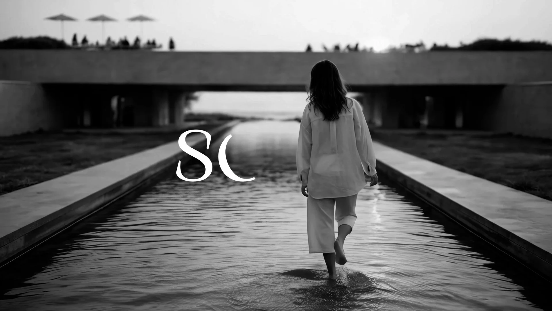 A person with long hair walking through water on a narrow path under a bridge at sunset or sunrise.