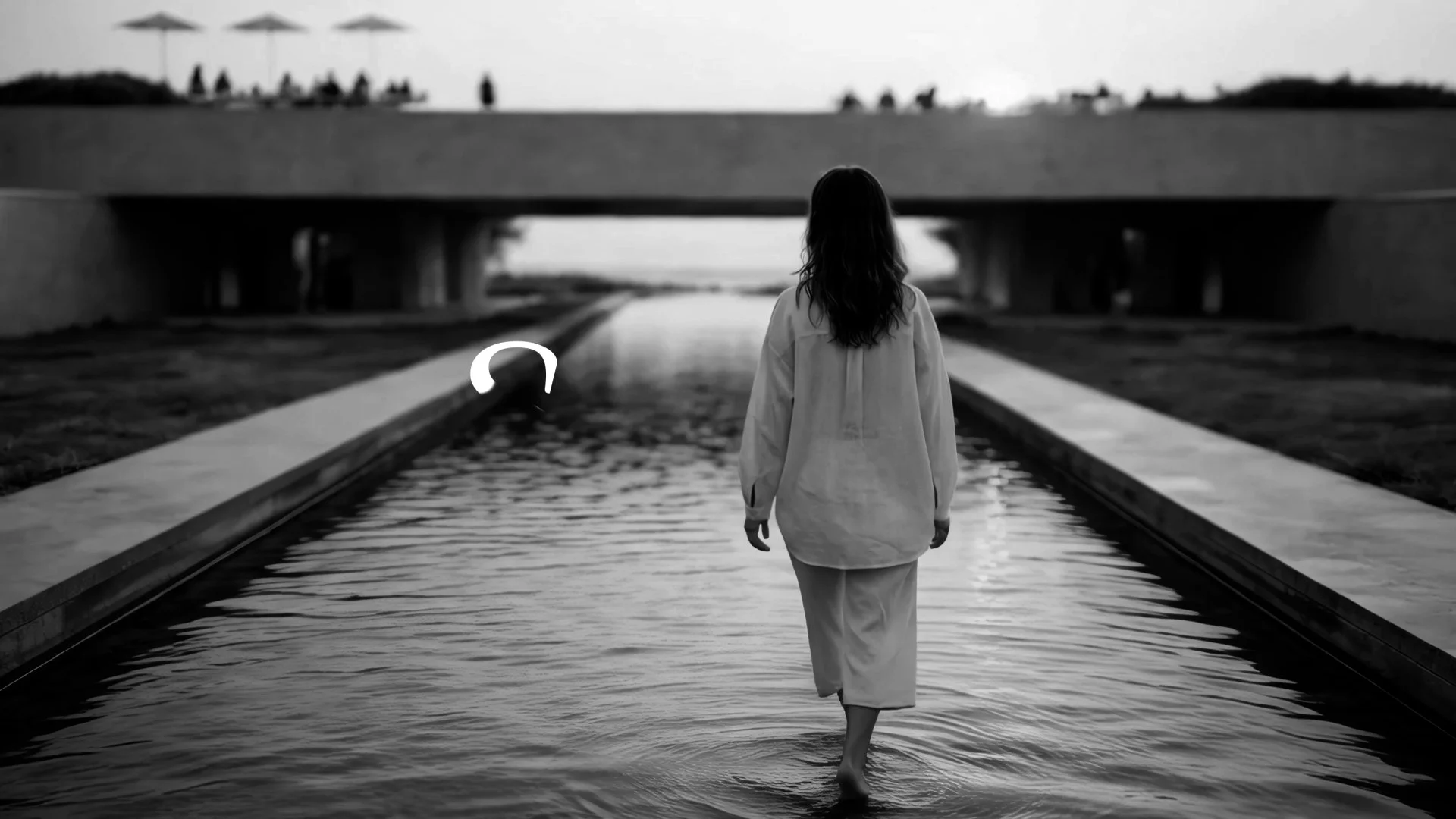 A woman walking barefoot through water on a rainy day, with a bridge and people in the background.