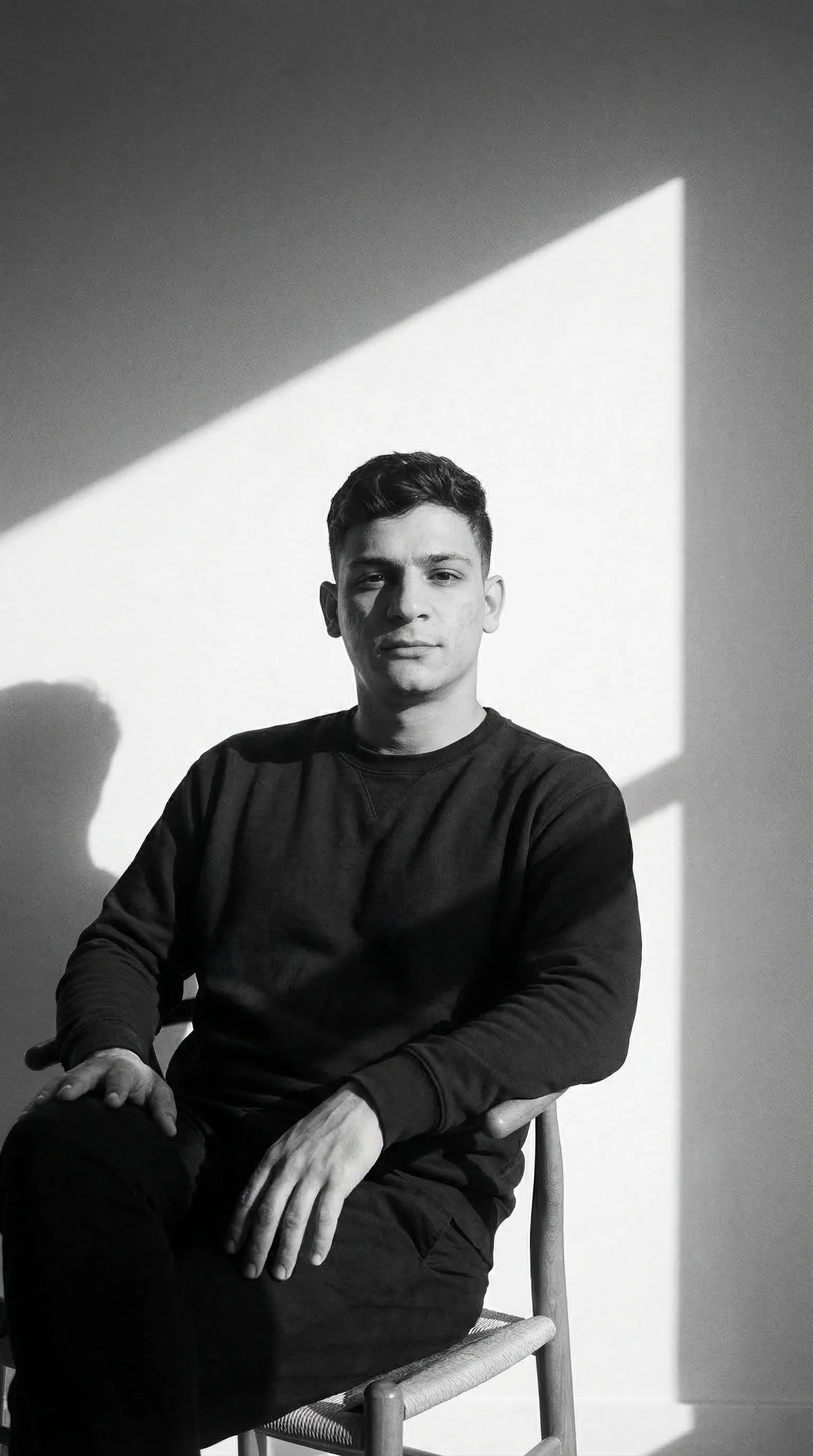 Black and white photo of a young man sitting on a wooden chair, with a large shadow of a window on the wall behind him.