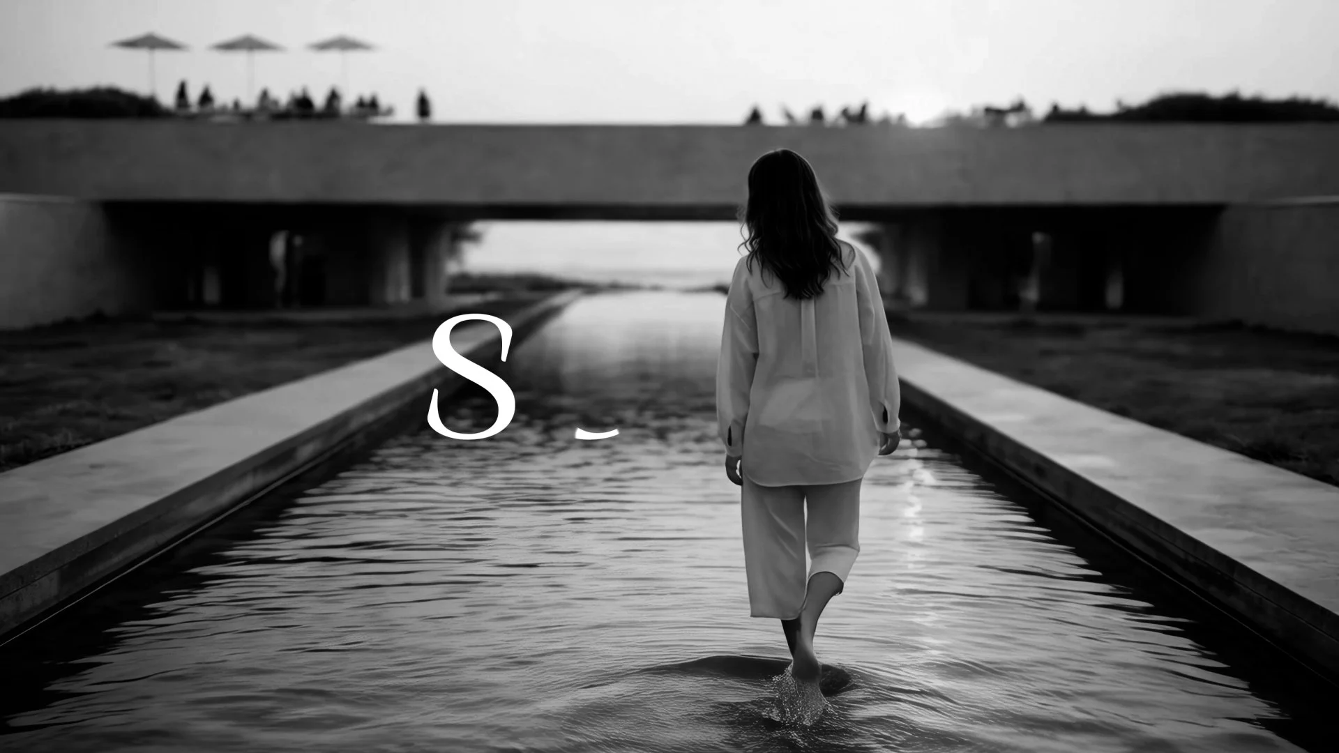 A person with long hair walking through a shallow water channel under a bridge during sunset, black and white photo.