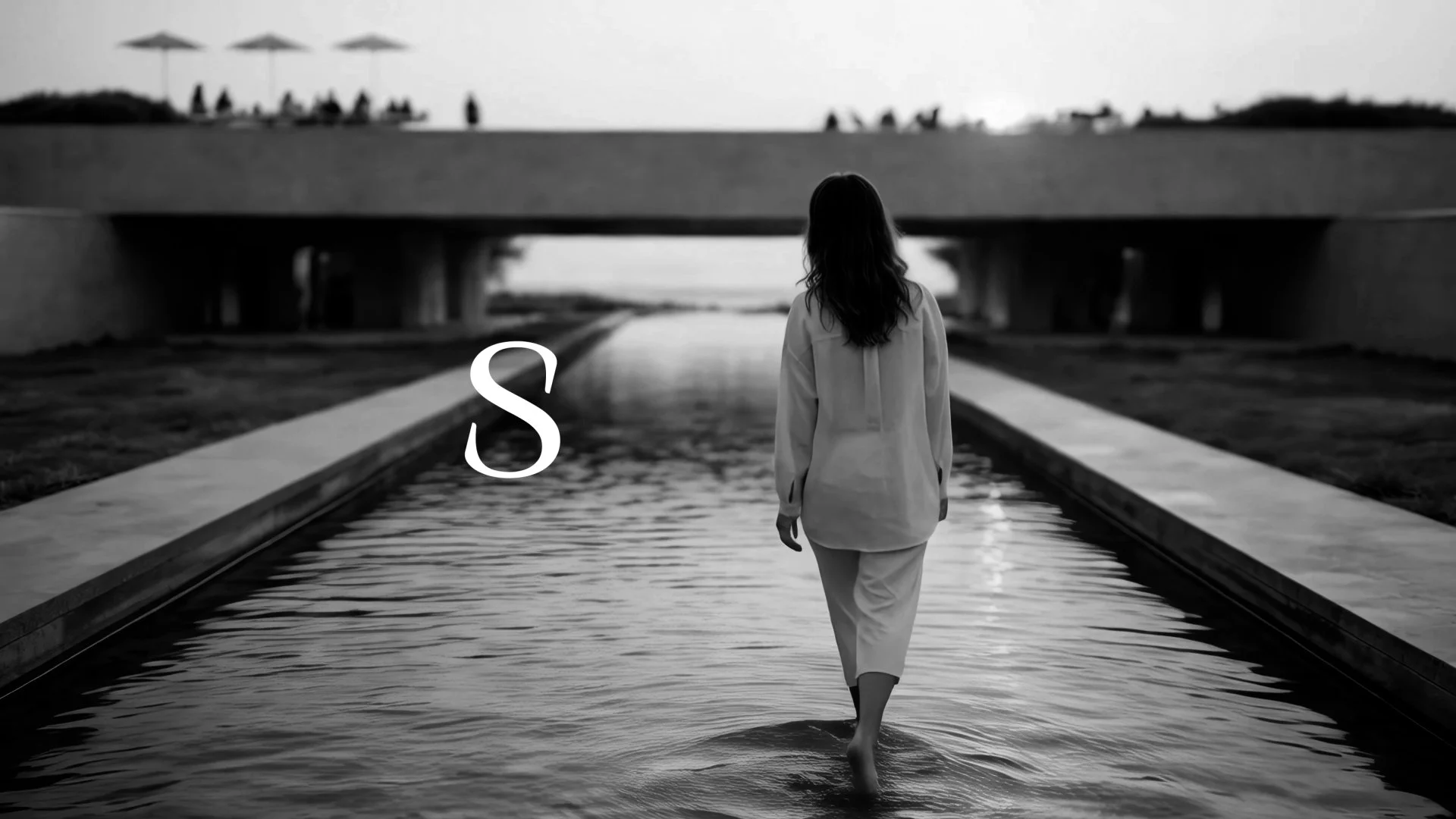 A woman in a light-colored outfit walking through a shallow water feature under a bridge at sunset with people and umbrellas on the bridge in the background. The photo is in black and white and features a large letter 'S' superimposed on the image.