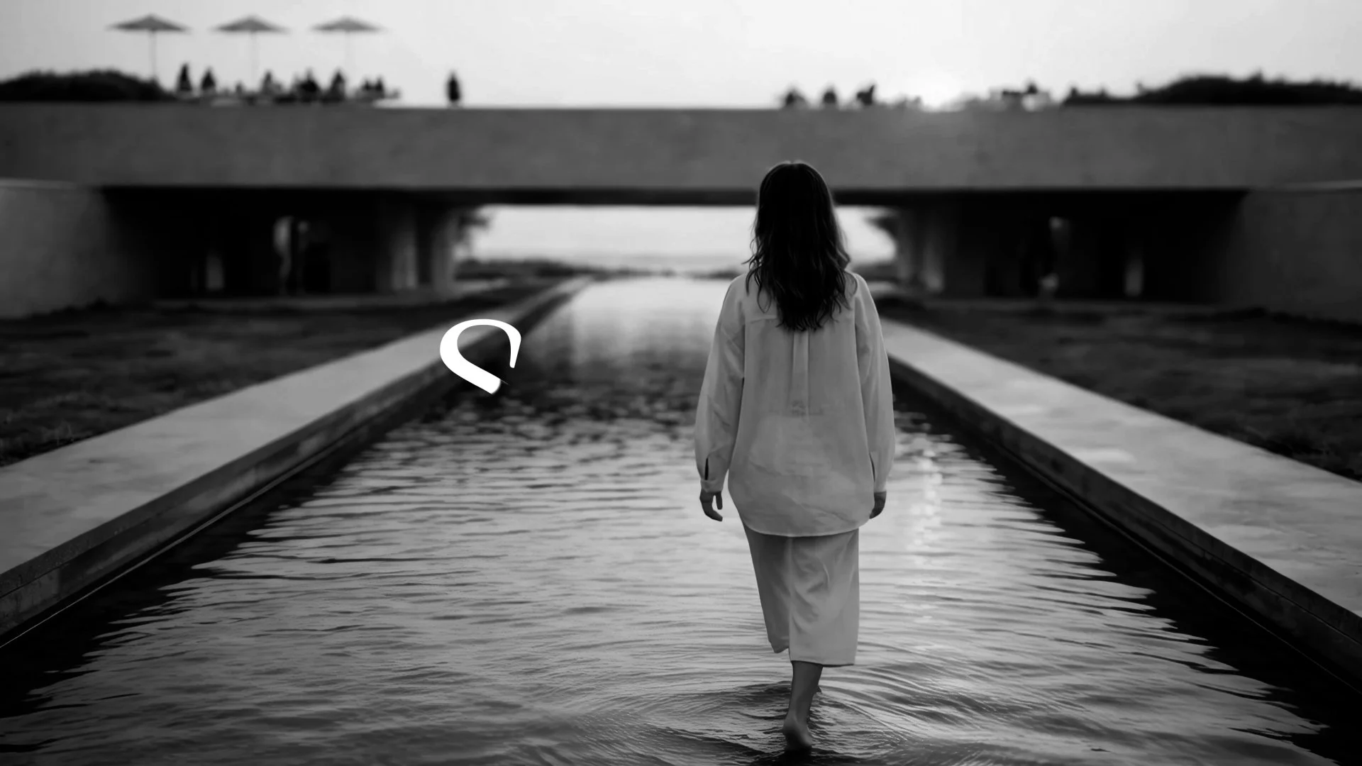 A woman is walking barefoot through a water-filled section of a pathway under a bridge, with the city skyline in the background, in black and white.