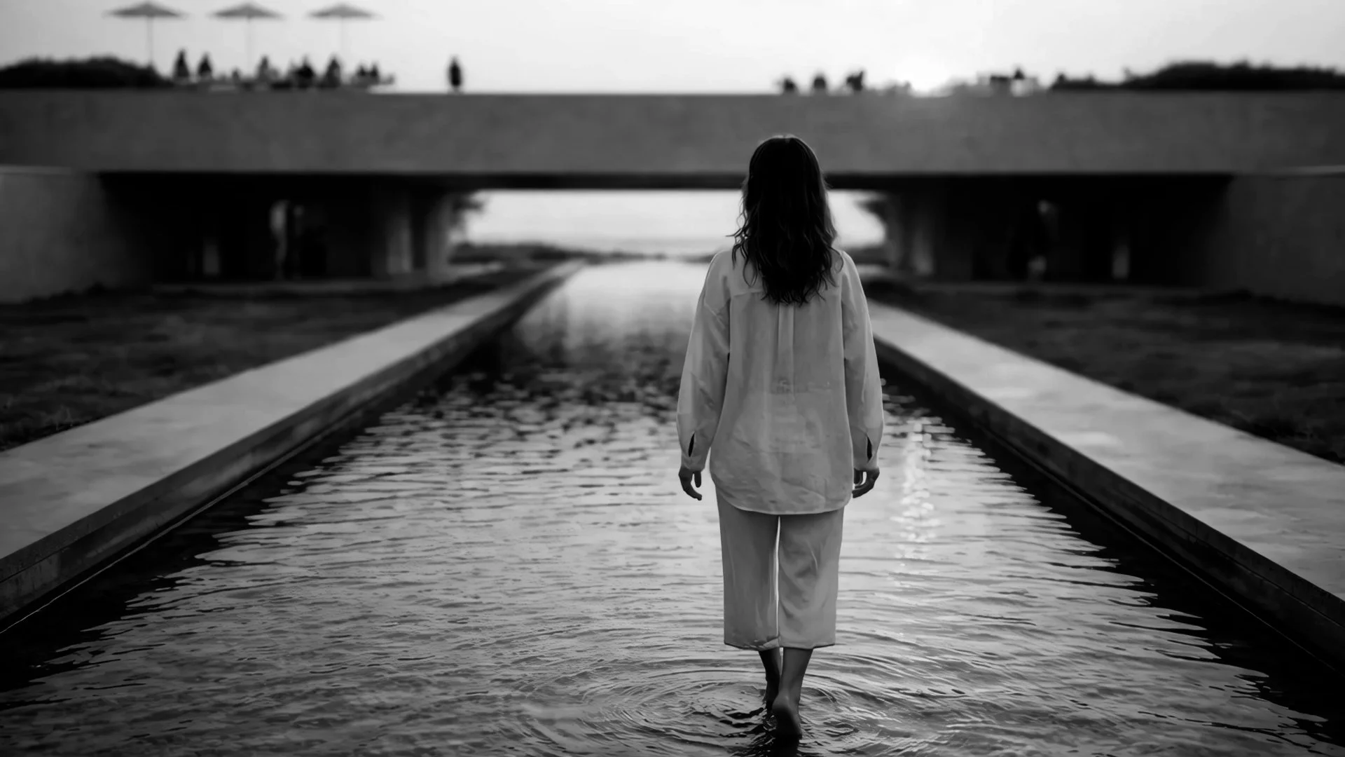 A woman with long hair, dressed in light-colored clothing, walks barefoot through a narrow water channel beneath a bridge during what appears to be end of day or early evening.