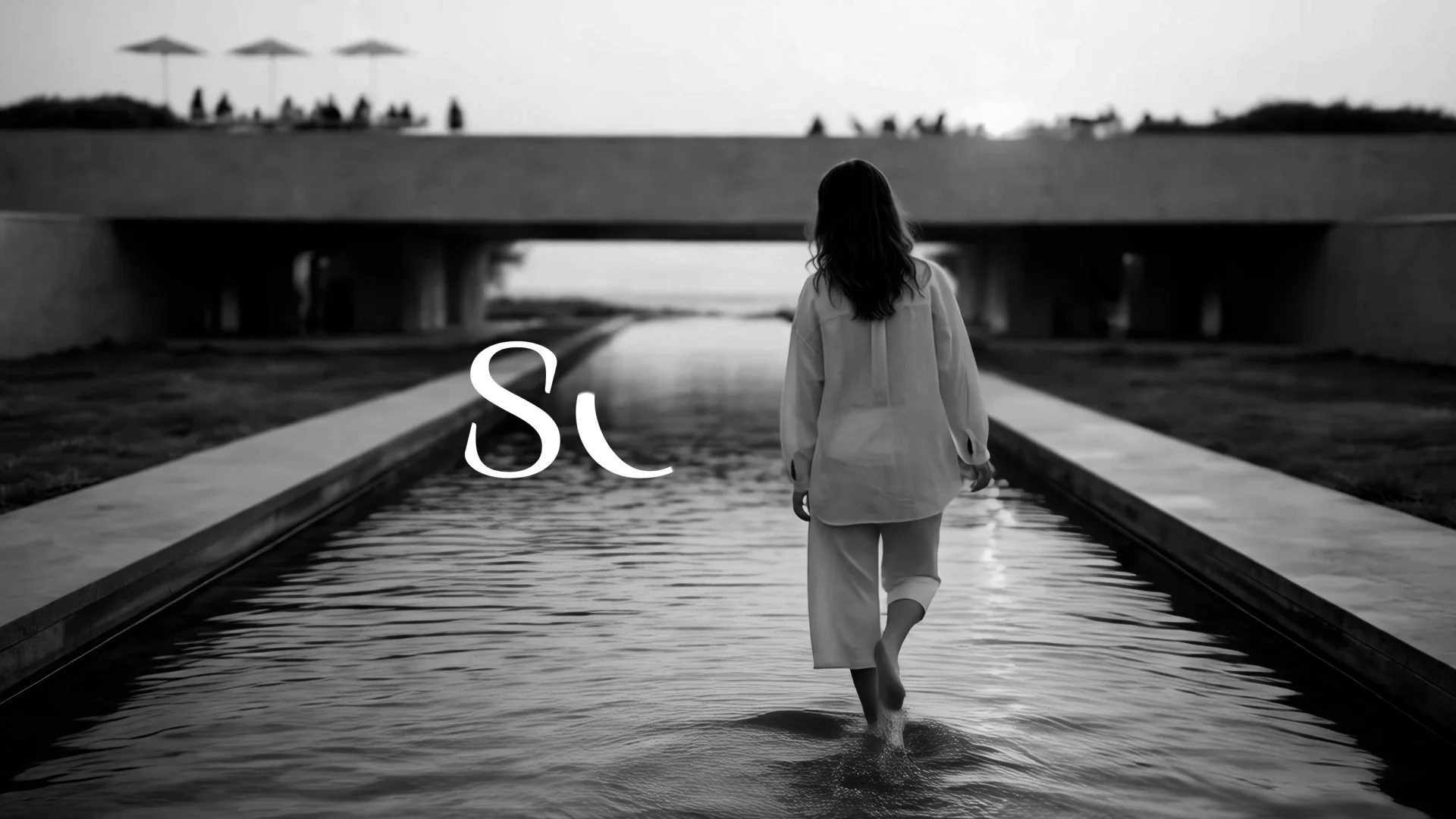 Black and white photo of a woman walking barefoot in a shallow water feature under a bridge, with trees and umbrellas in the background