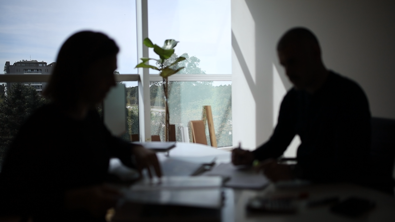 Silhouettes of two people sitting at a table indoors near a large window, with papers and documents in front of them, a potted plant, and a view of trees and buildings outside.