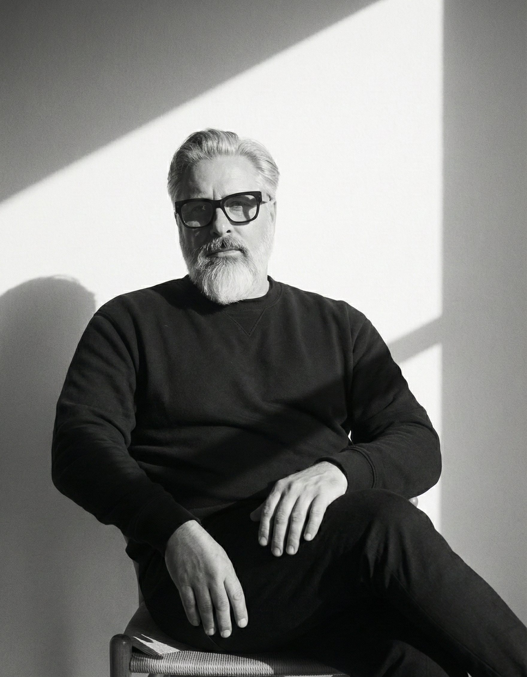 Black and white portrait of a man with glasses, a beard, and white hair, sitting on a chair against a wall with sunlight casting diagonal shadows.