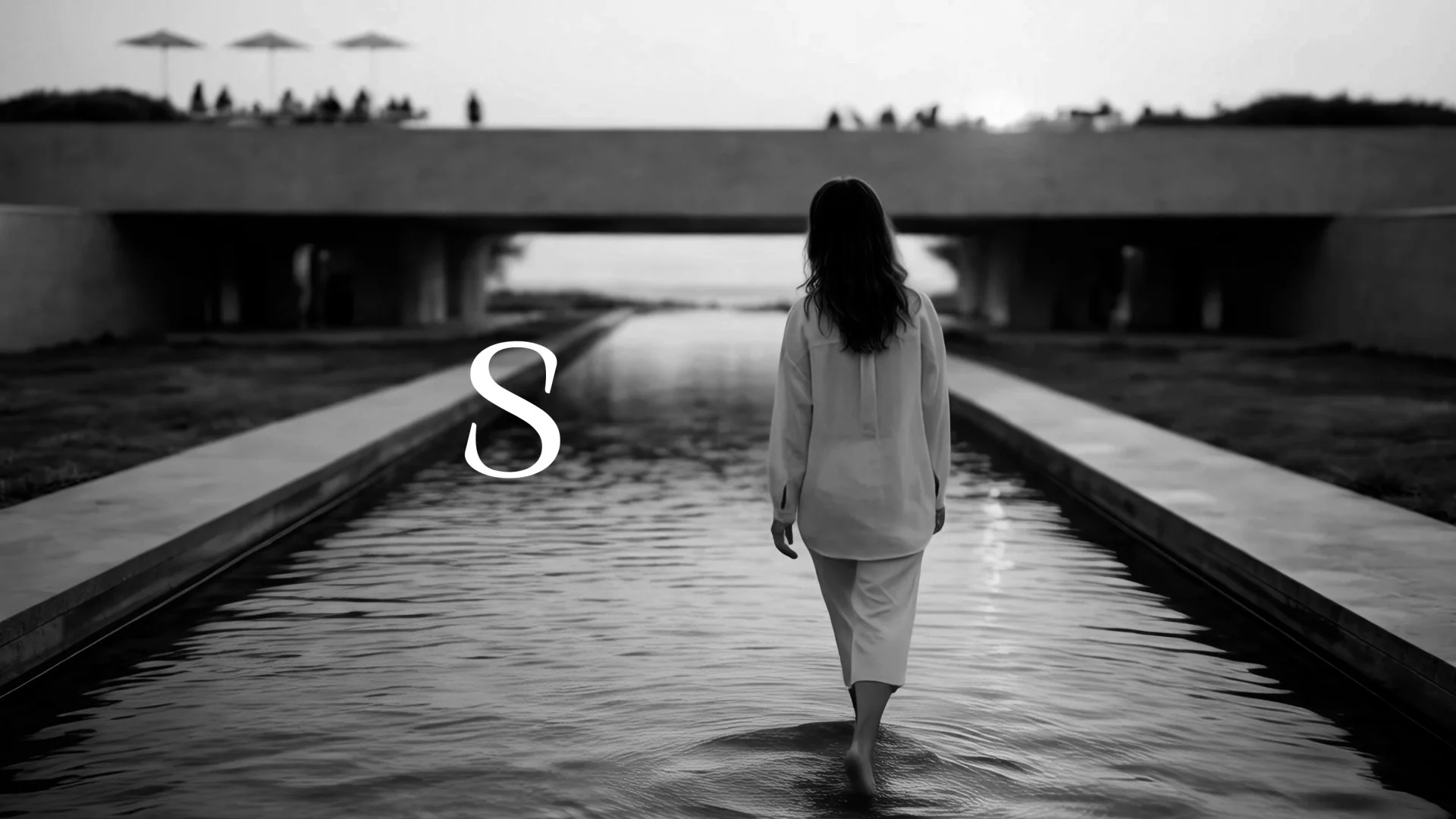 A black and white photo of a woman walking barefoot through a shallow water canal under a bridge, with people and umbrellas on a hill in the background.