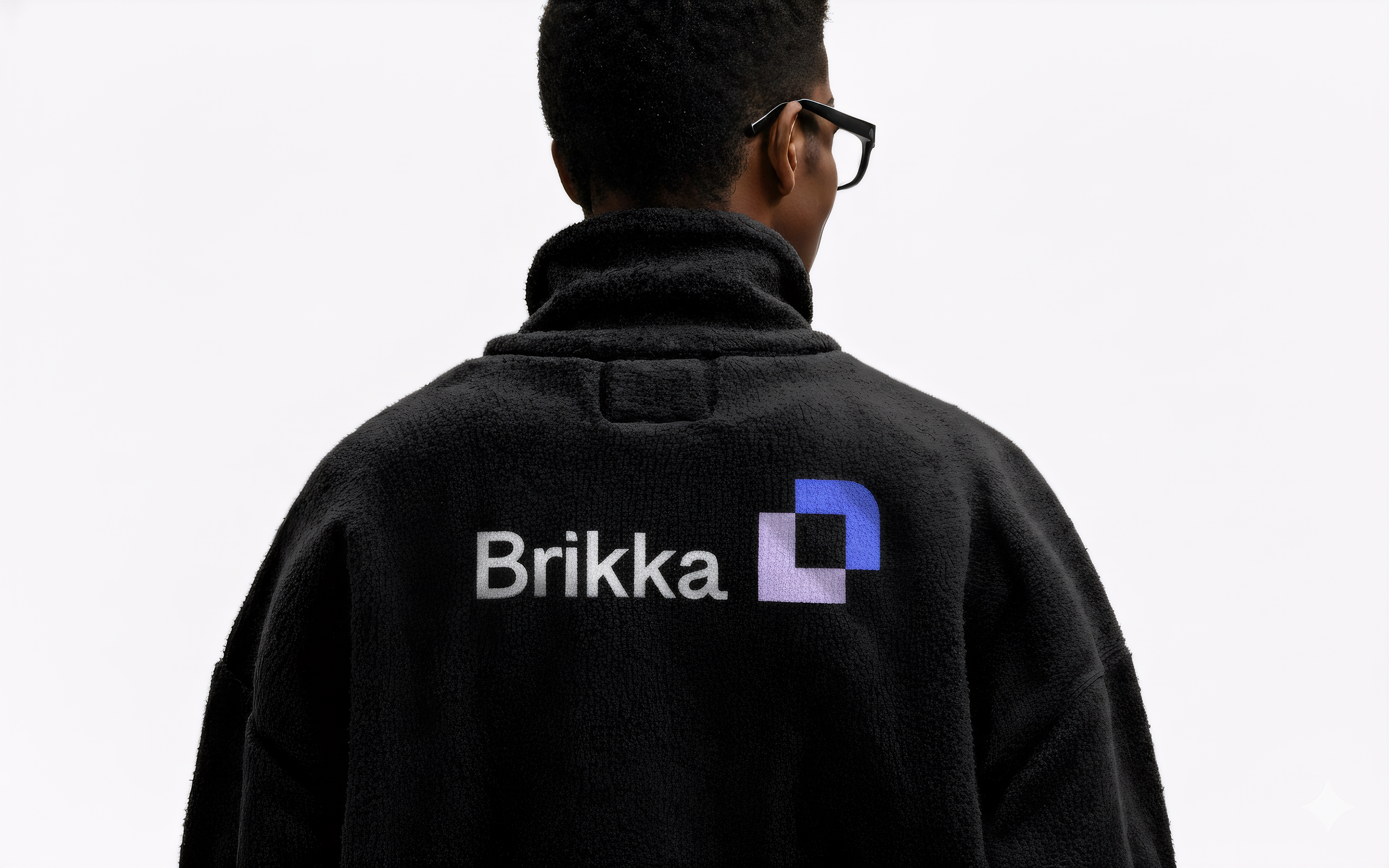 Back of a person wearing a black fleece jacket with the logo 'Brikka' and a geometric design in shades of blue and purple on the back. The person has short, curly hair, wears black-rimmed glasses, and is looking away from the camera against a plain white background.