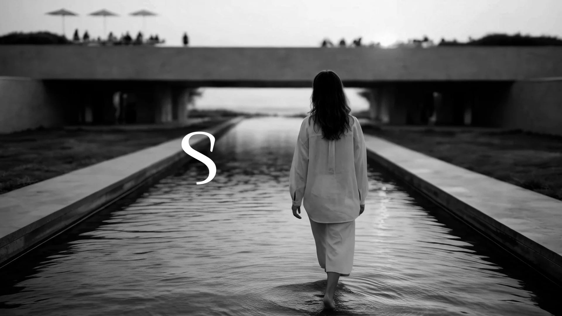 A woman with long hair wearing a long dress walking through water under an overpass at sunset, with people sitting on umbrellas on a hill in the background.
