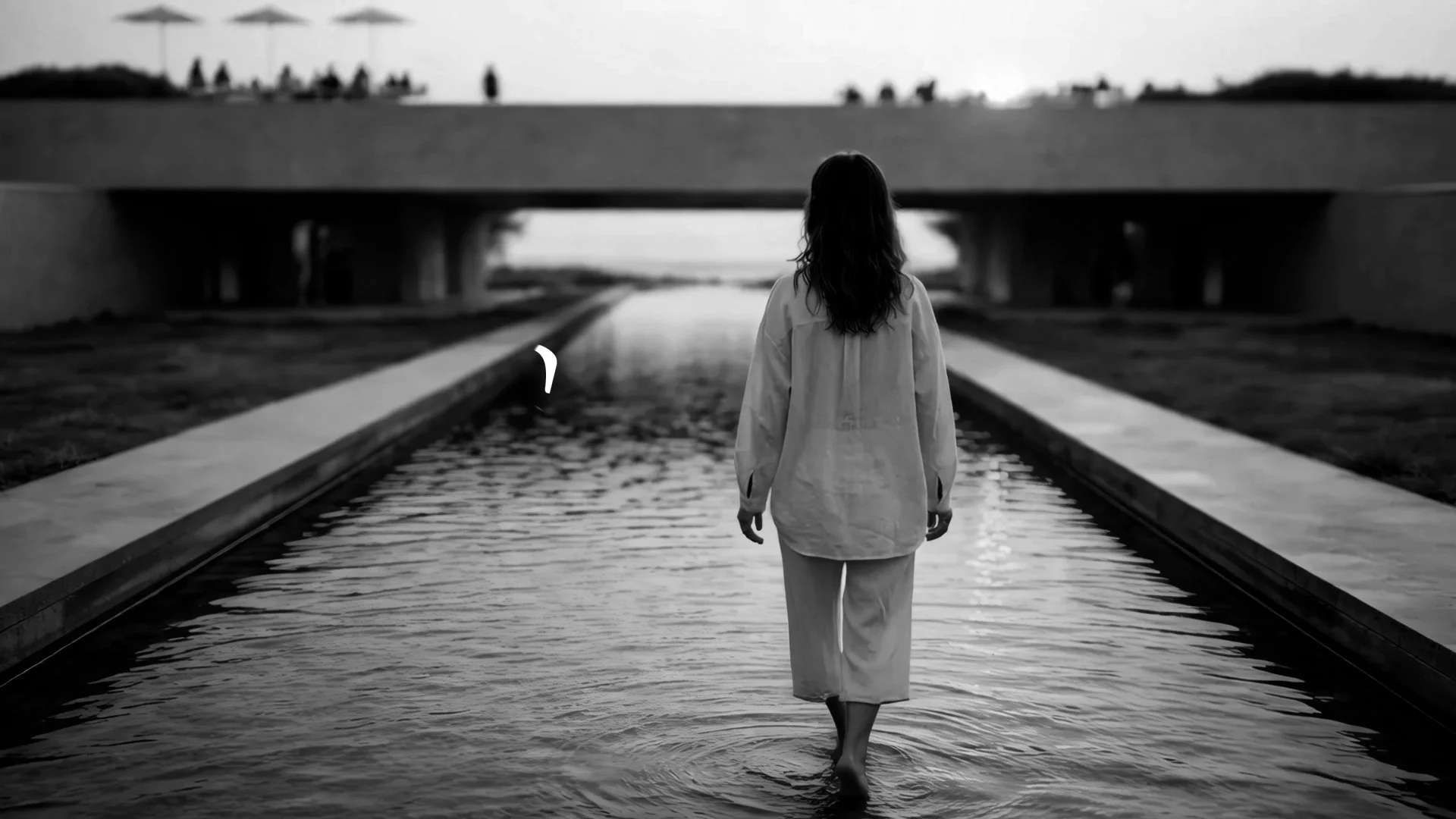 A woman walks barefoot through a shallow water canal under a bridge, with an overcast sky and distant figures on the bridge.