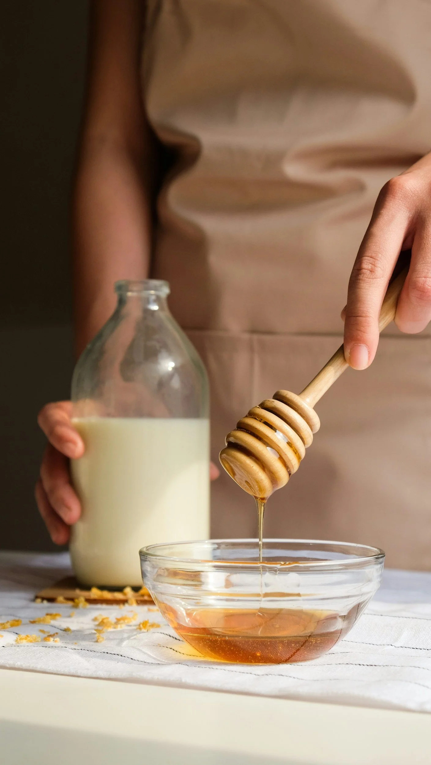 A person is holding a glass bottle of milk and a honey dipper with honey, dripping into a glass bowl of honey; the person is wearing a beige top.