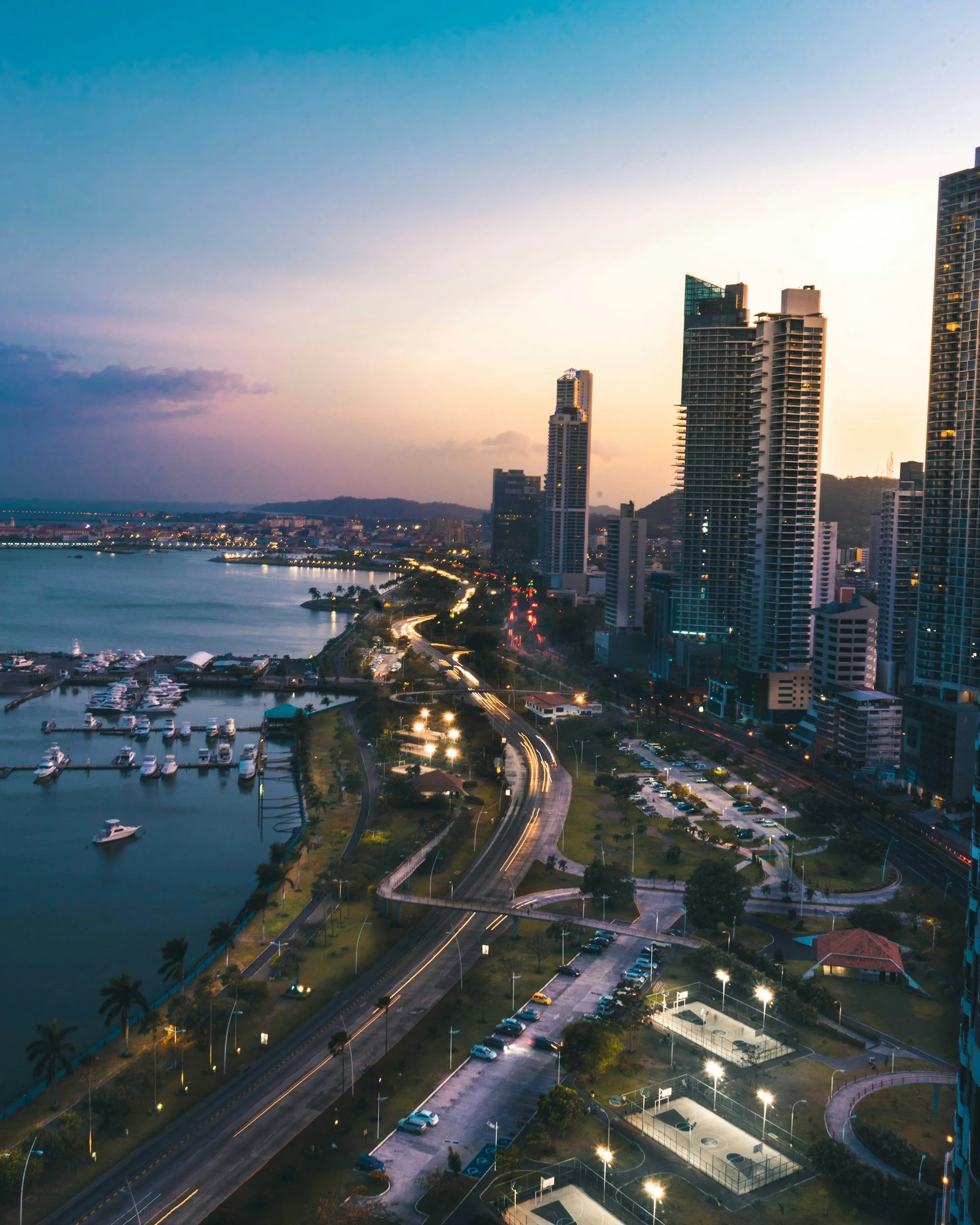 A cityscape at dusk with high-rise buildings, a marina with boats, and a park with courts and street lights.