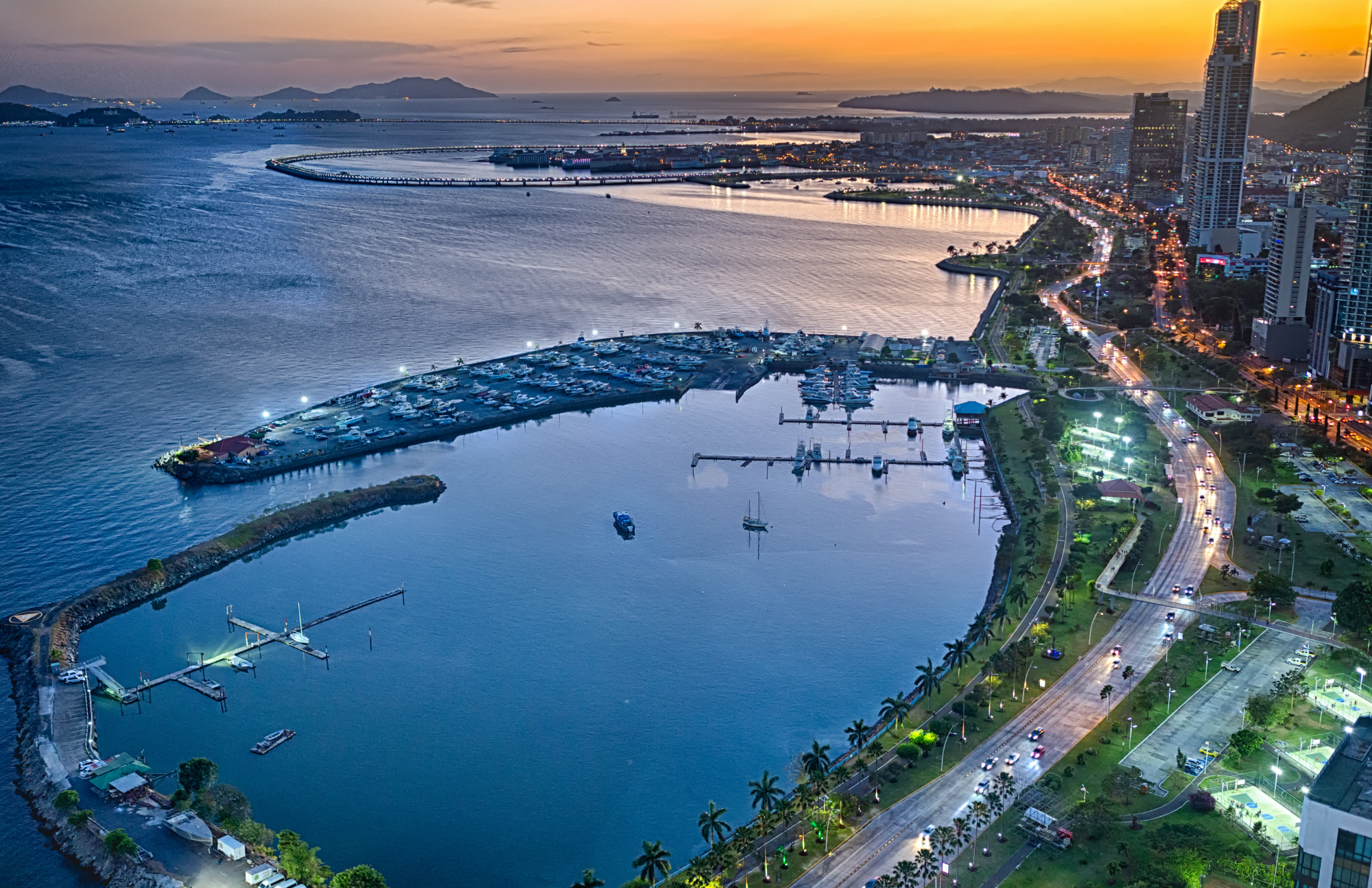 A city waterfront at sunset with buildings, a marina with boats, and a coastal road illuminated by streetlights.