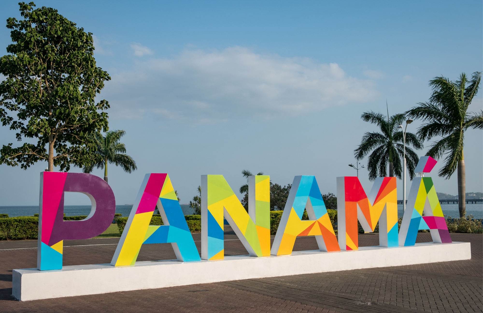 Colorful large letters spelling 'BAHAMAS' on a white rectangular base outdoors, with palm trees, a clear blue sky, and the ocean in the background.