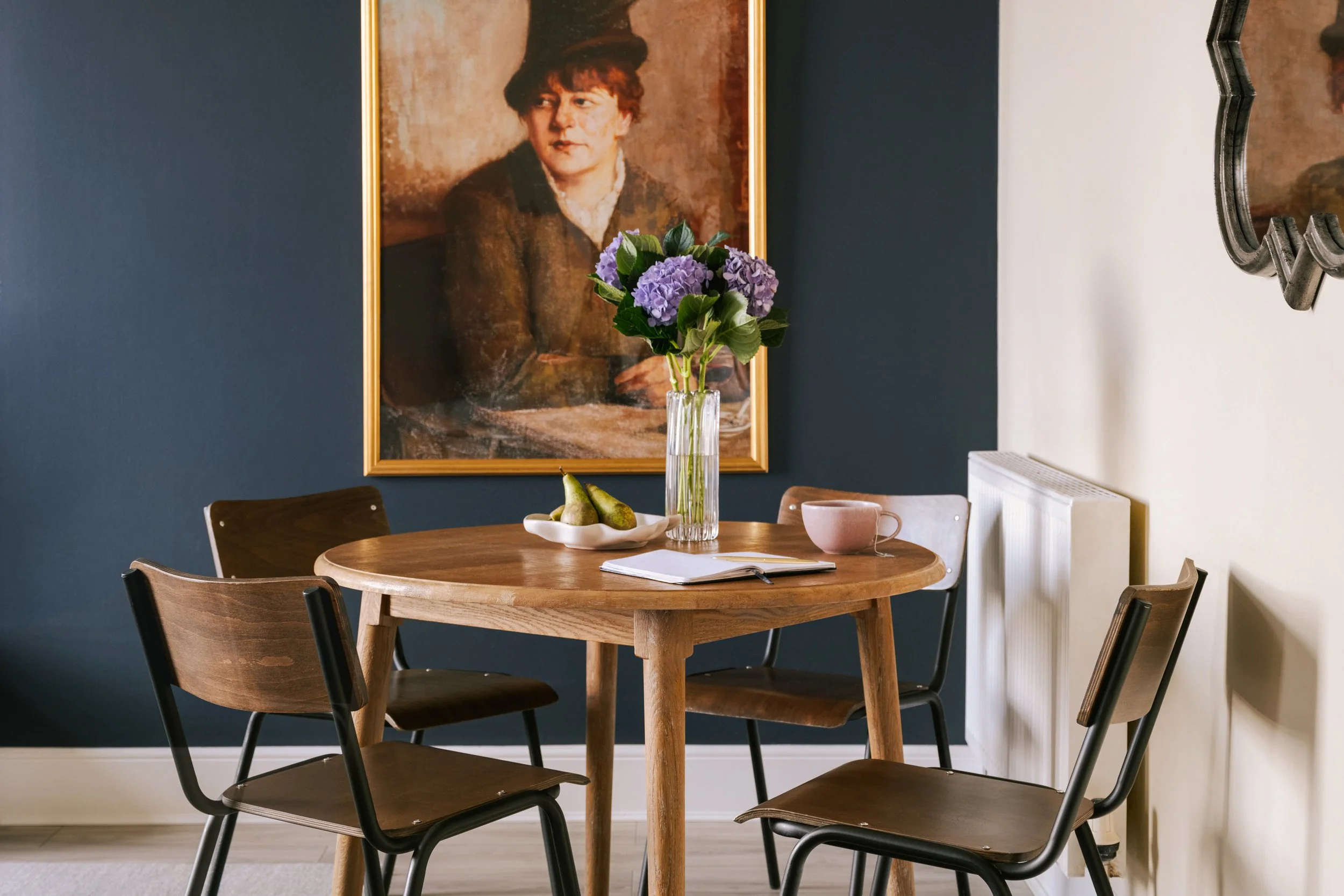 A wooden dining table with four chairs, a vase of purple flowers, a bowl of pears, a pink mug, and an open book, with a large portrait of a woman in vintage clothing hanging on a dark blue wall behind.