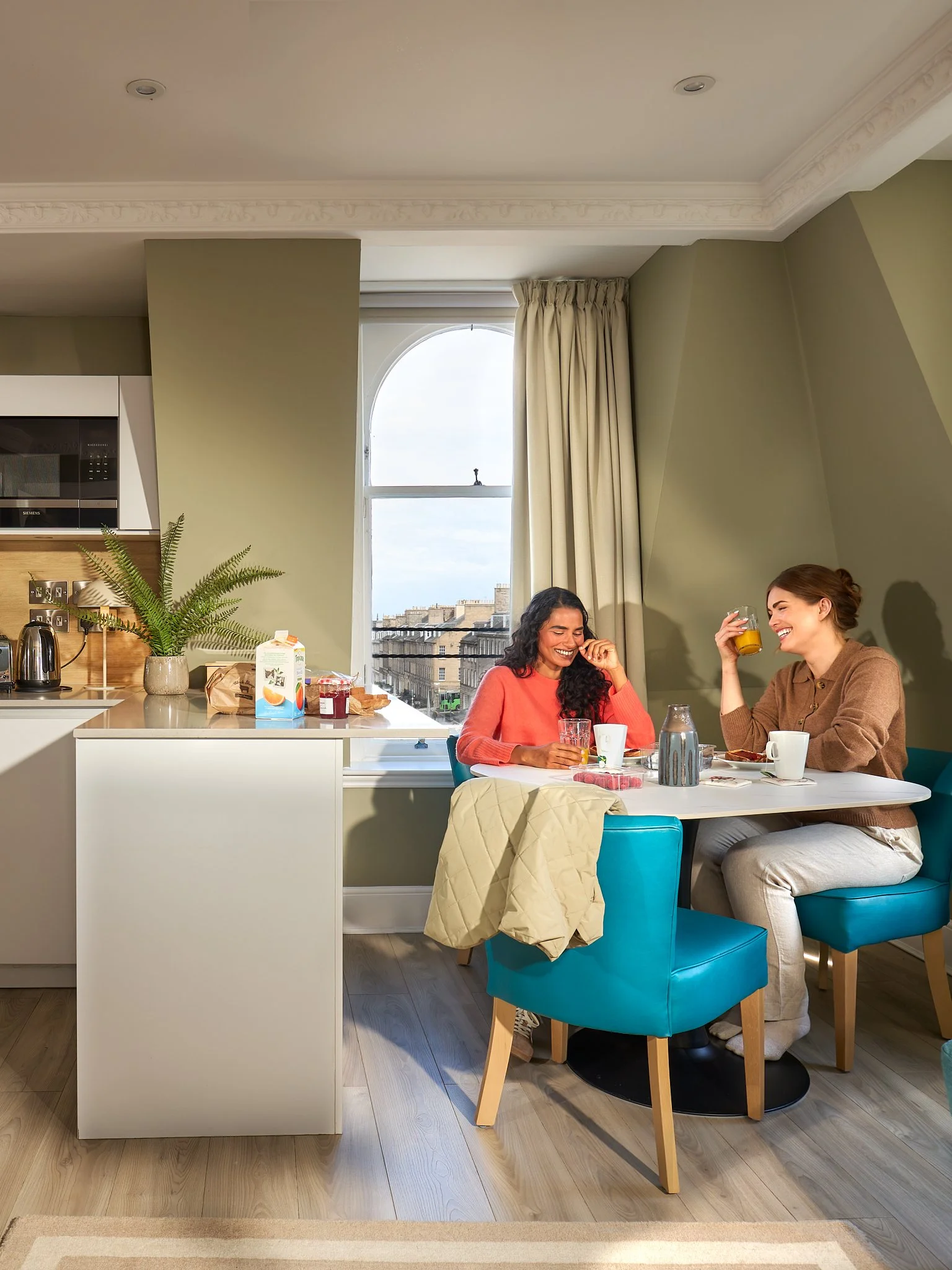 Two women sitting at a dining table, smiling and enjoying drinks in a well-lit kitchen or dining area with a window, curtains, and green walls.