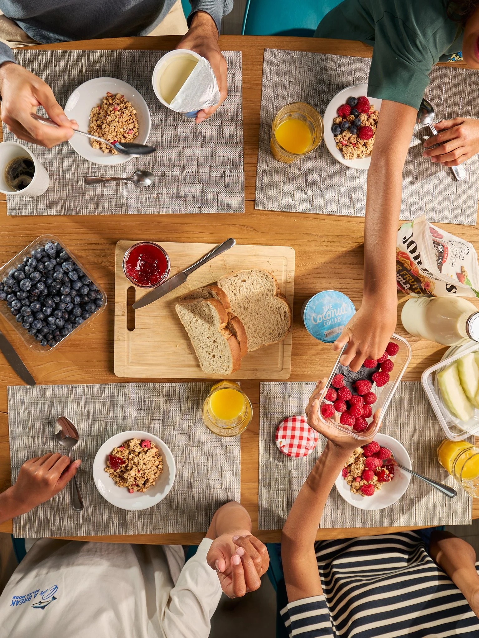 Top-down view of a breakfast table with bowls of cereal topped with berries, slices of bread, cups of coffee, orange juice, yogurt, and a container of blueberries and raspberries, with hands reaching for food.