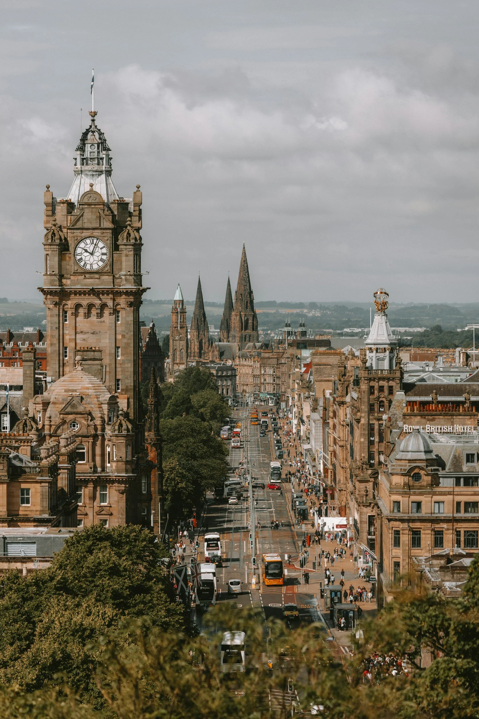 Cityscape of a historic European city with clock towers, medieval spires, and busy streets filled with cars and pedestrians under a cloudy sky.