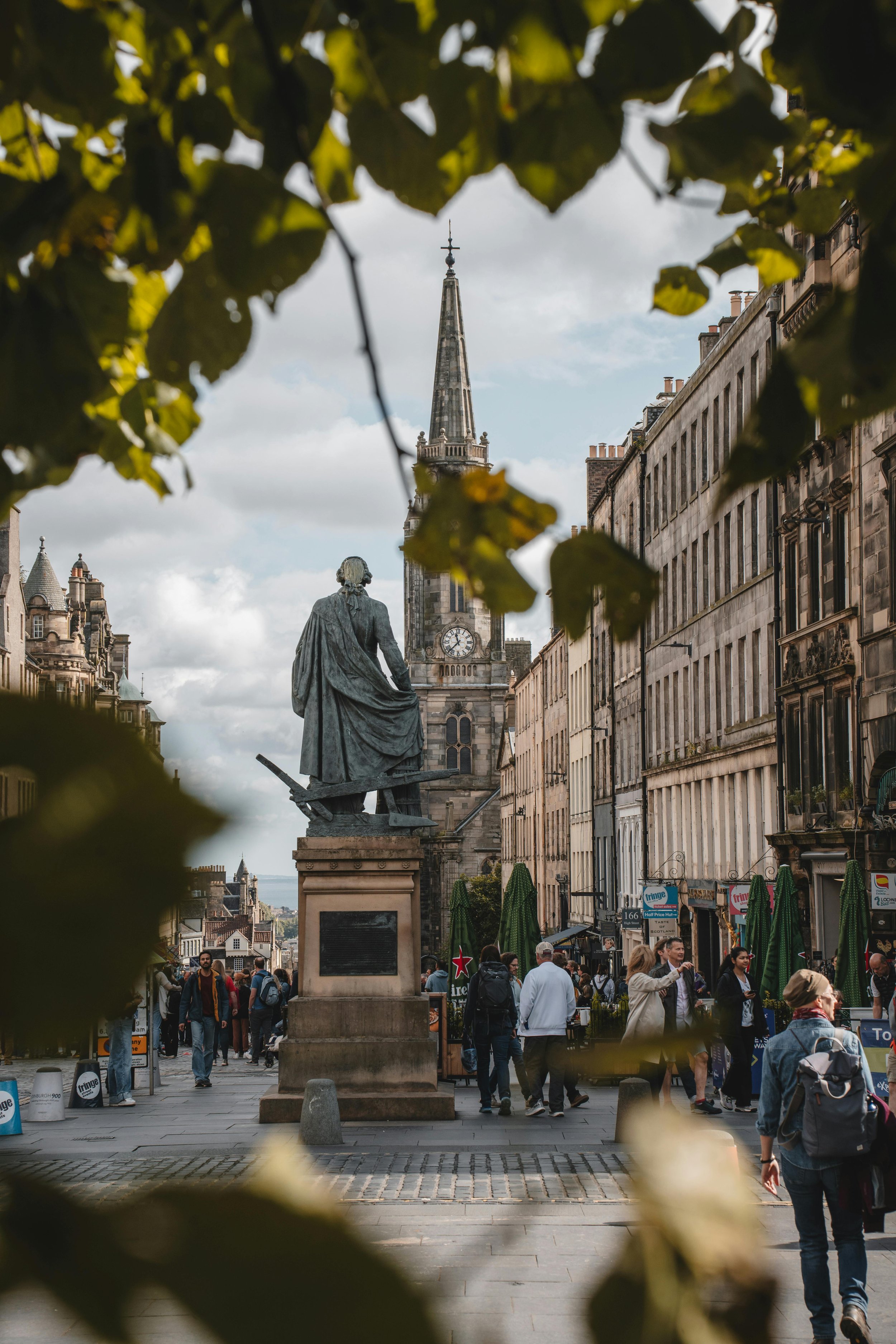 Street scene in Edinburgh, Scotland, with a statue in the foreground, historic buildings lining the street, and a church steeple in the background, viewed through foliage.