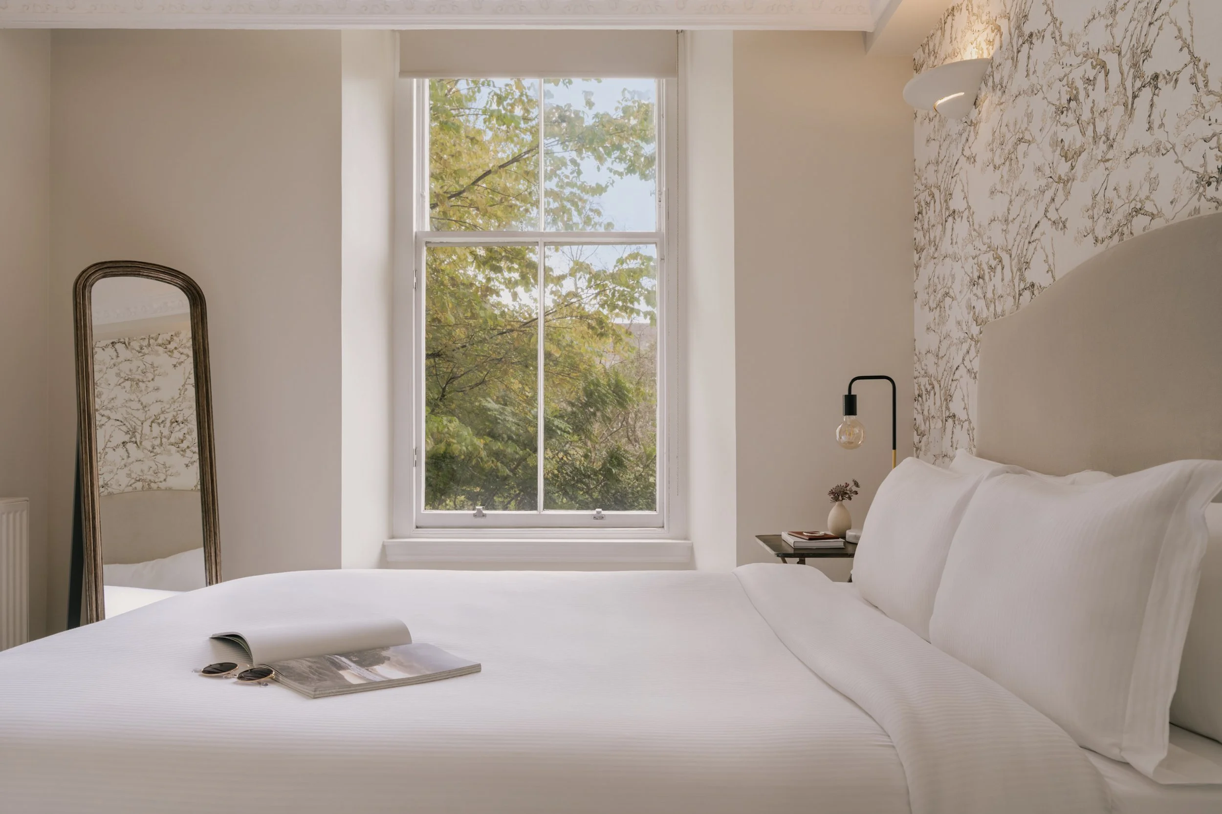 A bedroom with a large window showing trees outside. The bed has white bedding with an open magazine and sunglasses on it. A bedside table with a vase of flowers and a lamp is visible.