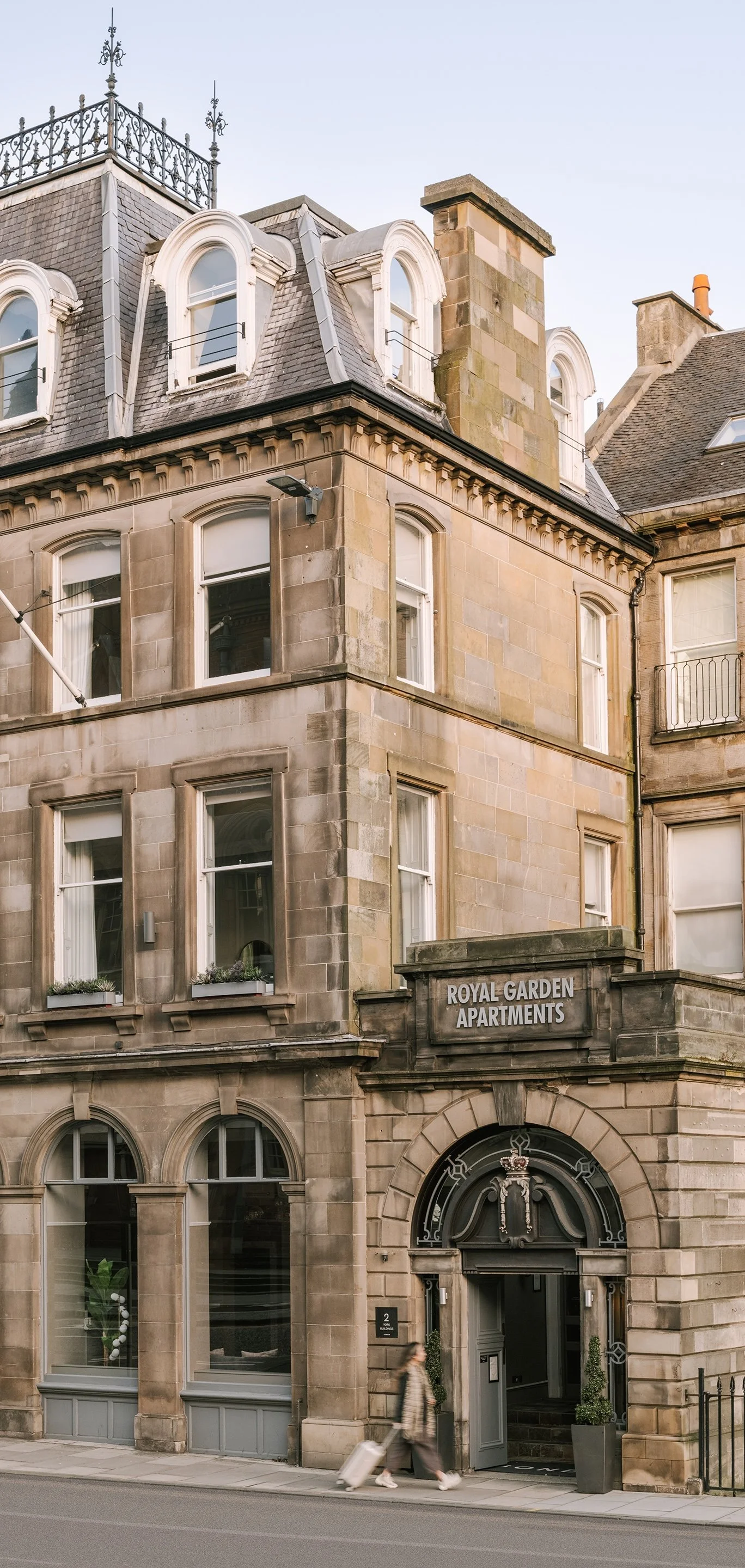 A historic stone building labeled "Royal Garden Apartments" with arched windows and a decorative entrance, with a person walking past pulling a suitcase.