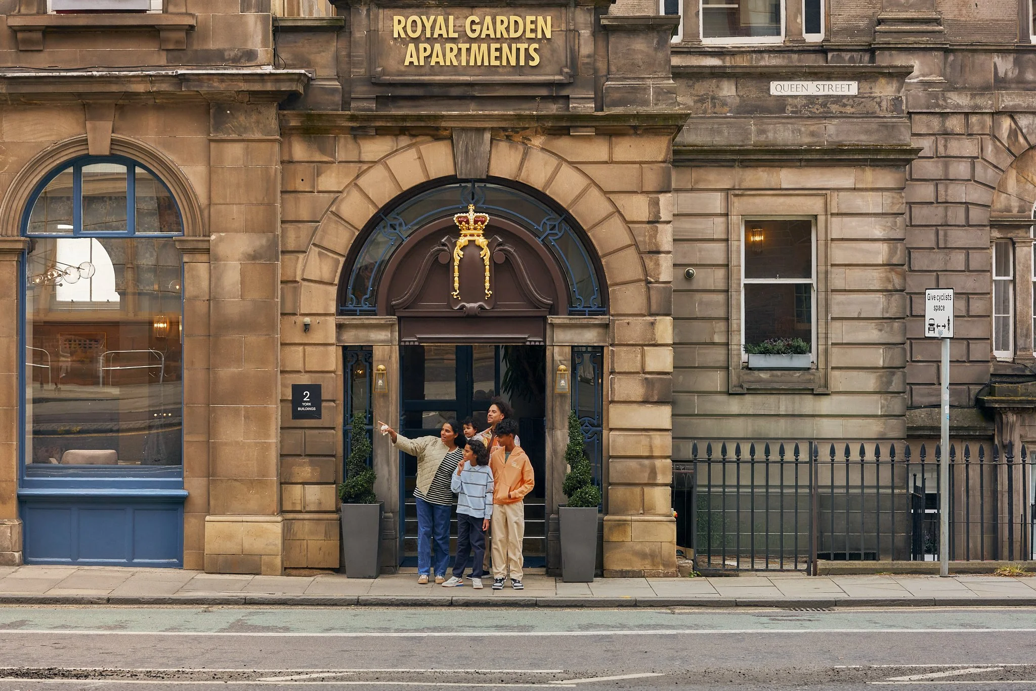 A group of four people, including a woman, two children, and a man, standing outside a historic building with a sign that reads "Royal Garden Apartments" at the entrance. They are taking a selfie or photo together.