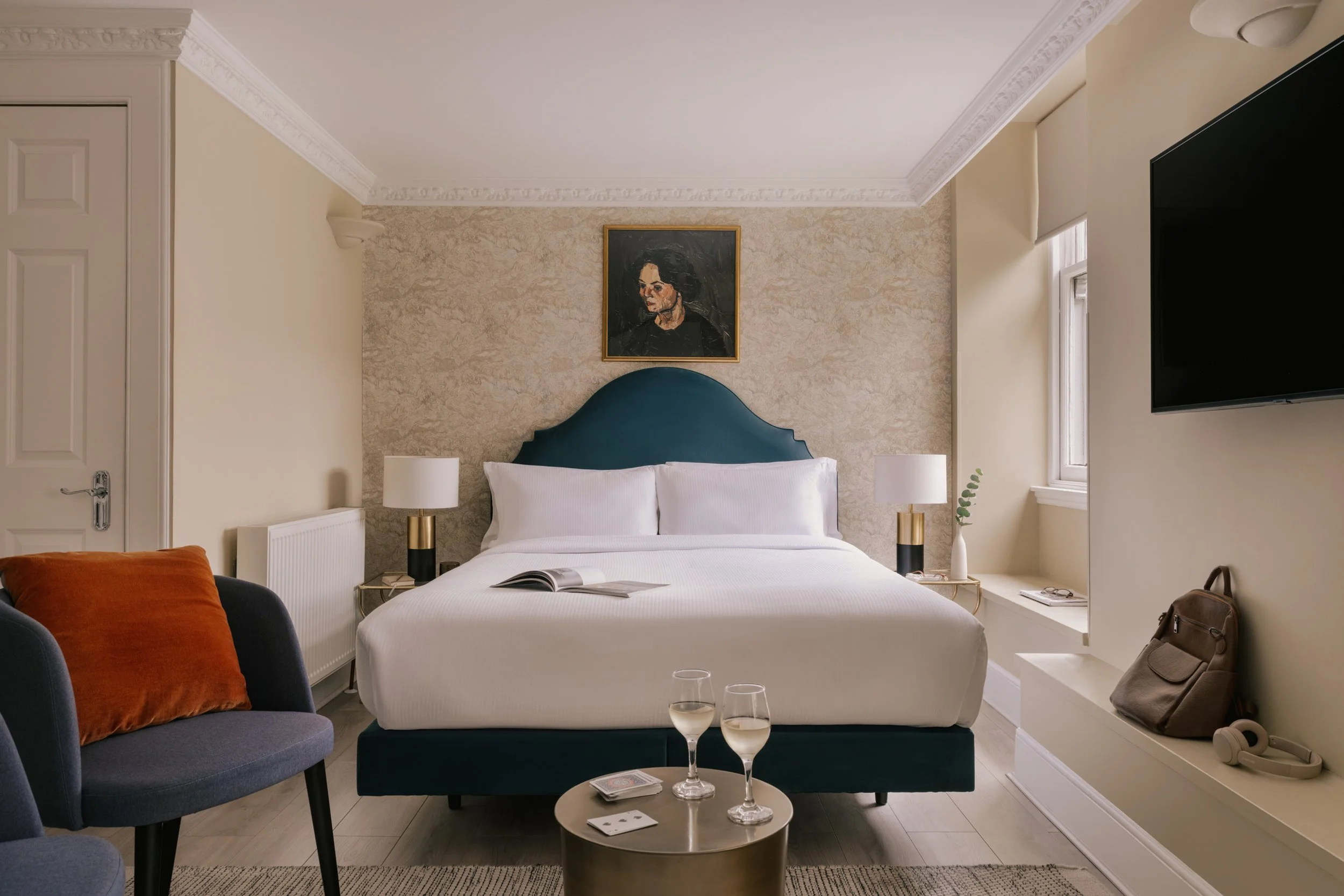 A hotel bedroom with a bed that has a white headboard and white bedding, two bedside tables with white lamps, a framed portrait above the bed, a small table with two glasses of white wine, a black armchair with an orange pillow, a TV on the wall, and a window with a blind.