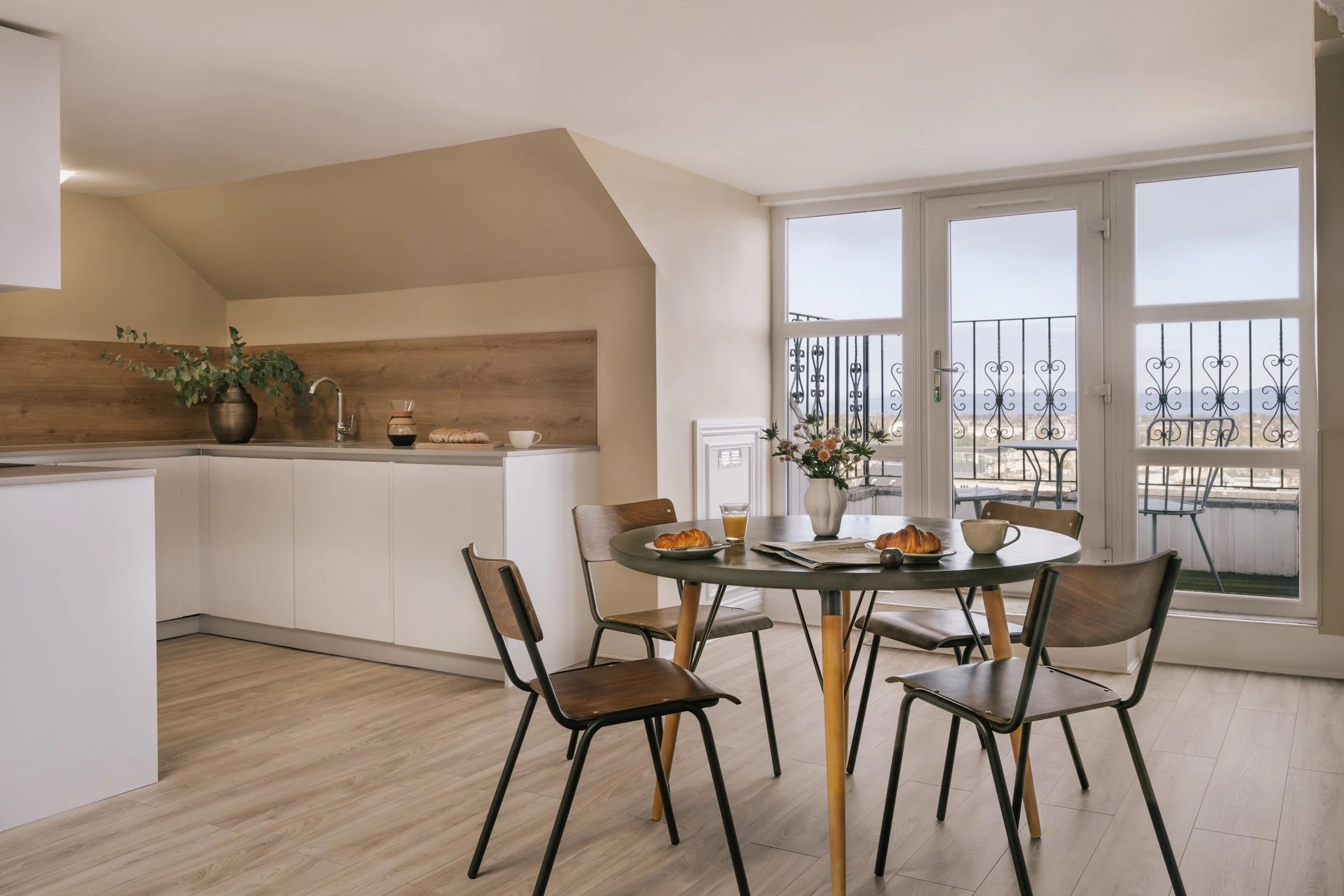 A modern kitchen with a dining area featuring a round table set with croissants, a newspaper, a mug, and a glass of orange juice. Large windows with iron balconies let in natural light, and there are chairs around the table and a plant on the countertop.