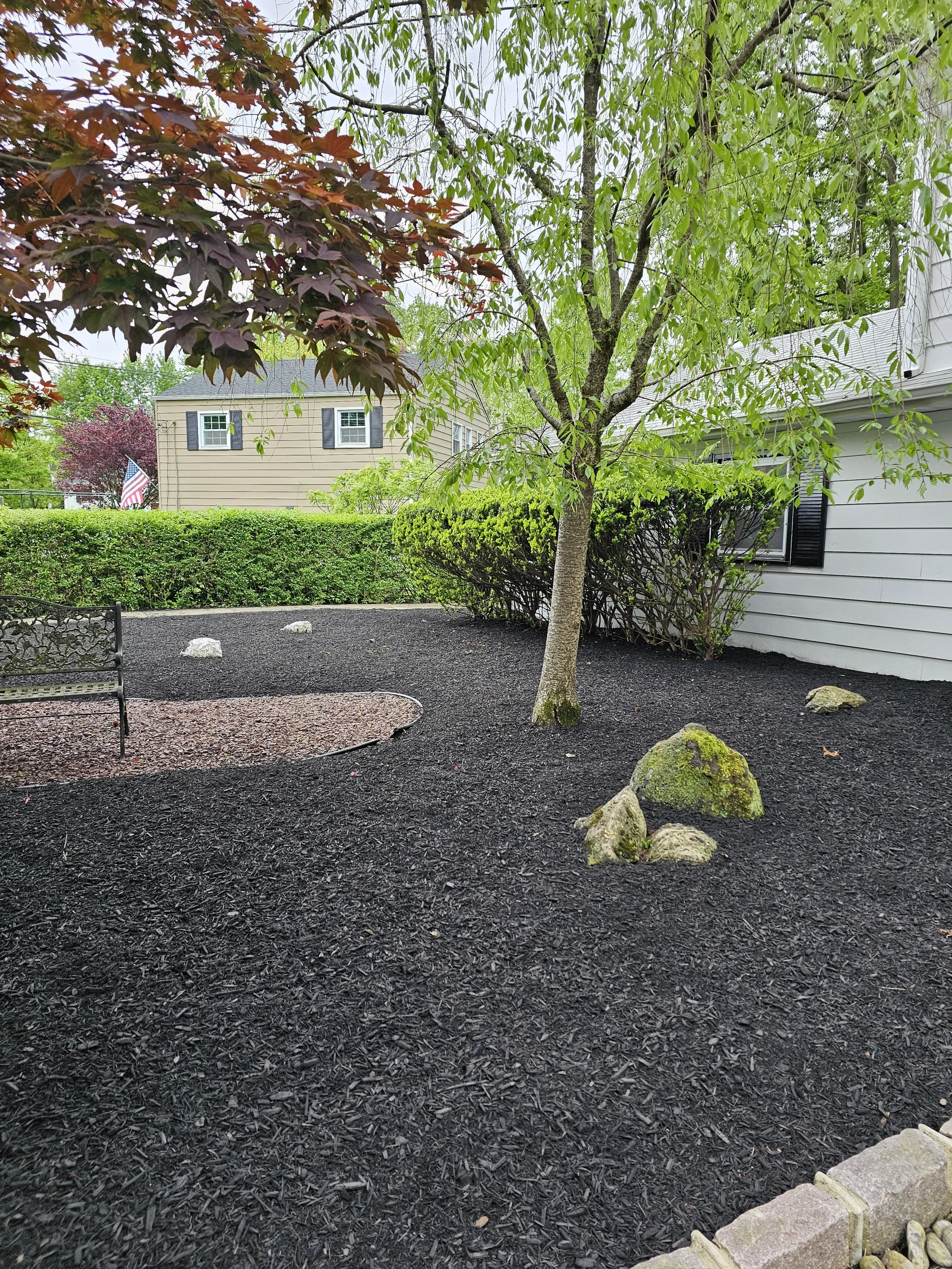 A backyard garden with a tree, mulch ground, a stone border, a bench, bushes, and a house with beige siding.