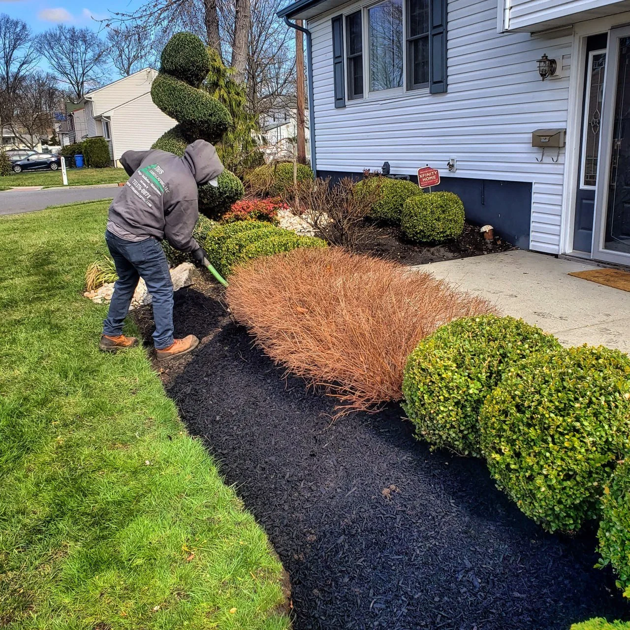 A person in a gray jacket and brown boots planting or tending to a freshly laid black mulch bed in front of a house with white siding and blue shutters.