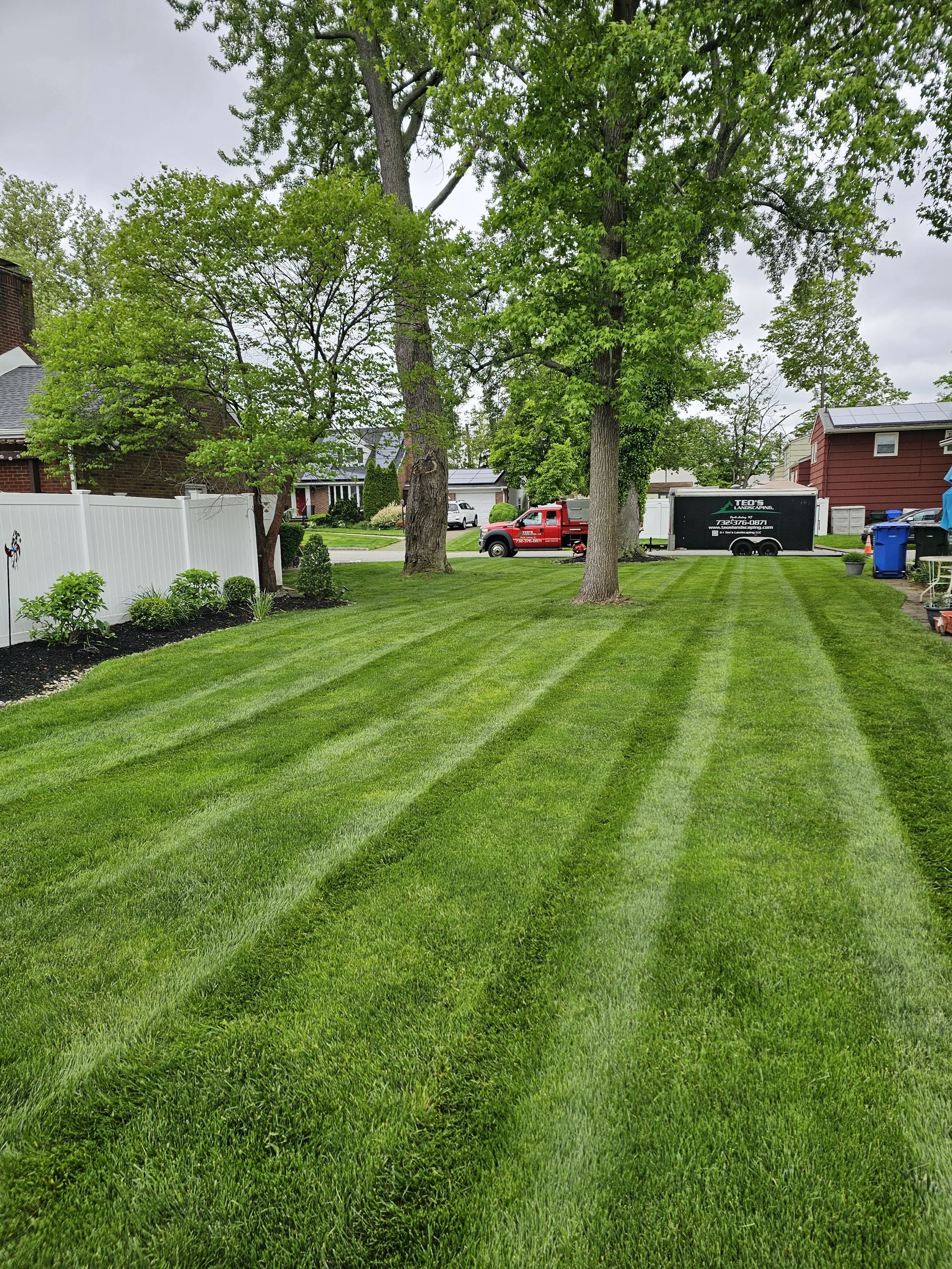 A well-manicured front yard with lush green grass, neatly cut with visible mowing lines. There are large trees and shrubs along the edges, and a white fence on the left side. In the background, there are houses, a red truck, and a black trailer parked on the street.
