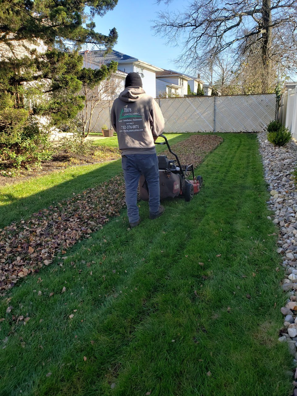 A person wearing a grey hoodie and black beanie operating a commercial leaf blower on a grassy lawn in a backyard, with trees, shrubs, and houses in the background.