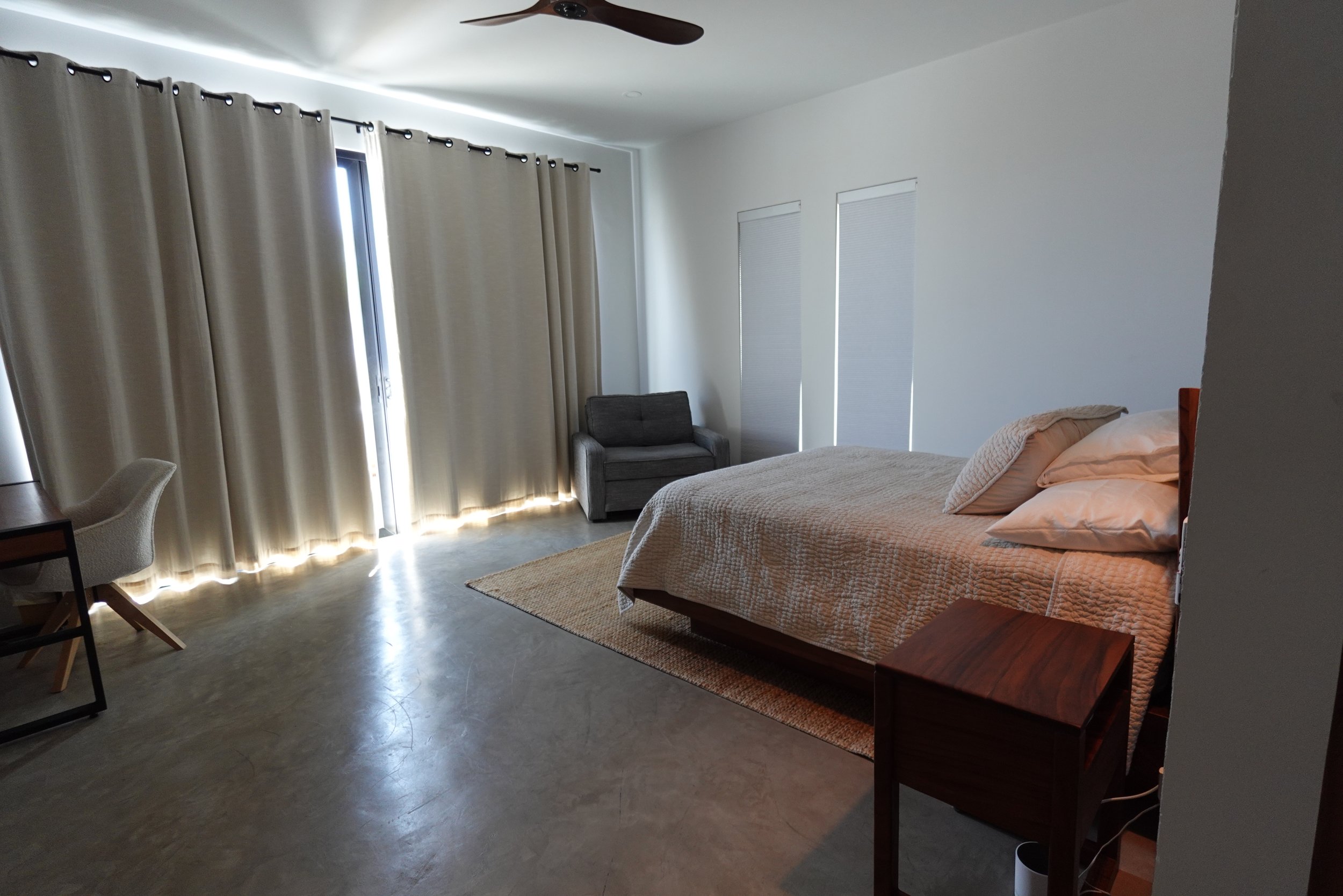 A bedroom with a bed having beige bedding and pillows on the right side, a gray armchair in front of tall windows with beige curtains on the left, a ceiling fan above, and a beige rug on the floor.