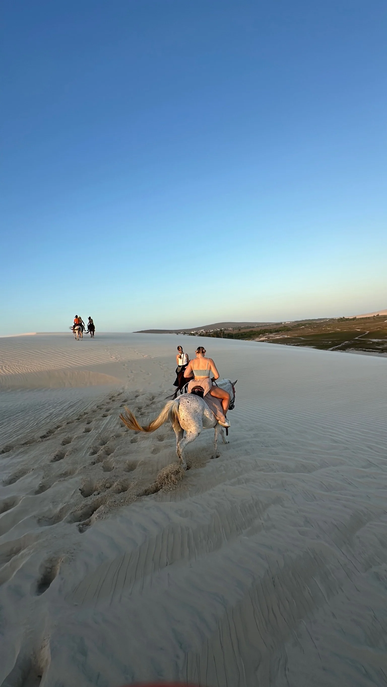 People riding horses on a sandy landscape with a clear blue sky and distant hills.