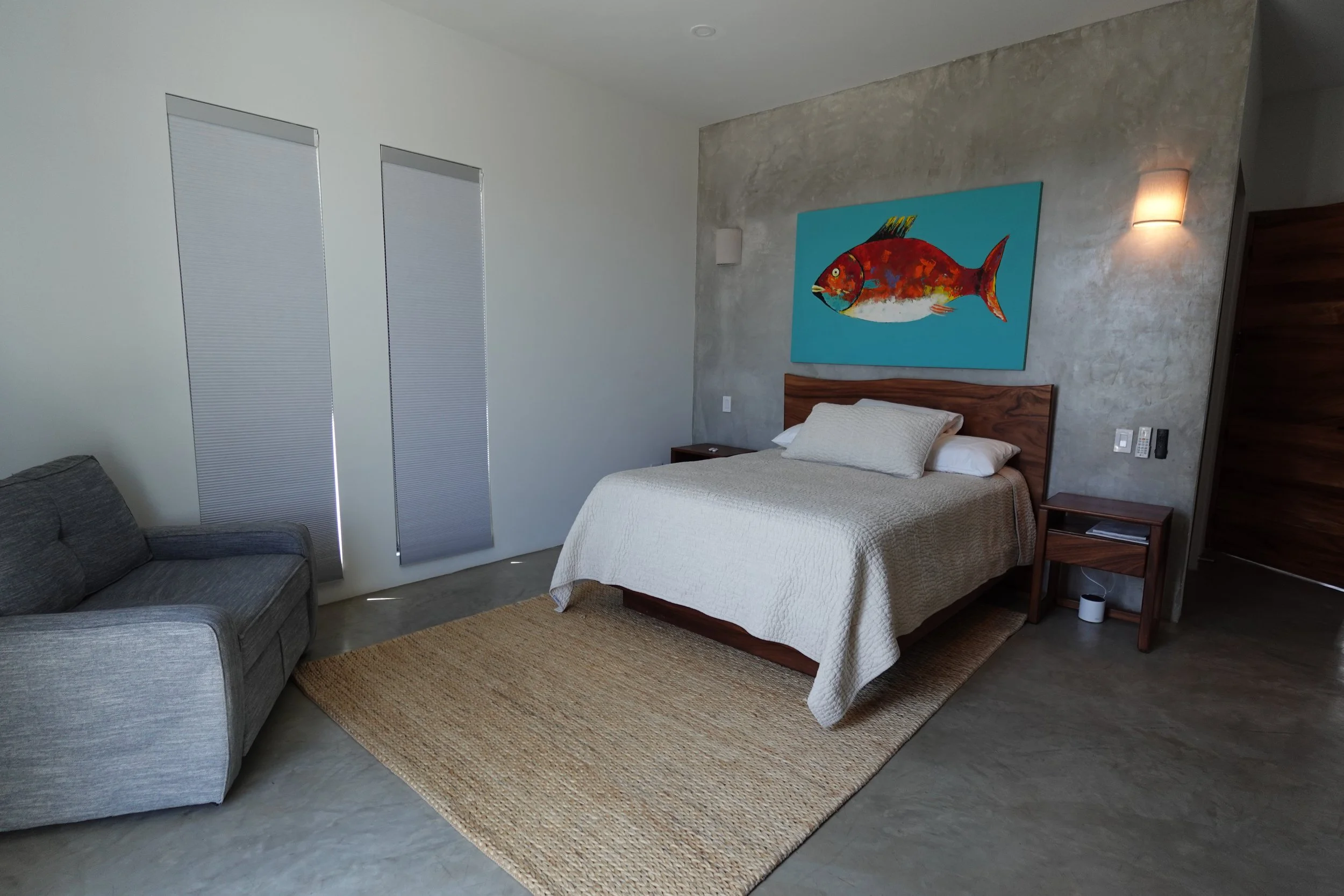A modern bedroom with a large bed, a wooden headboard, and a colorful fish painting on the concrete wall behind the bed. There is a nightstand with a remote control and a small lamp, and a beige rug on the concrete floor.