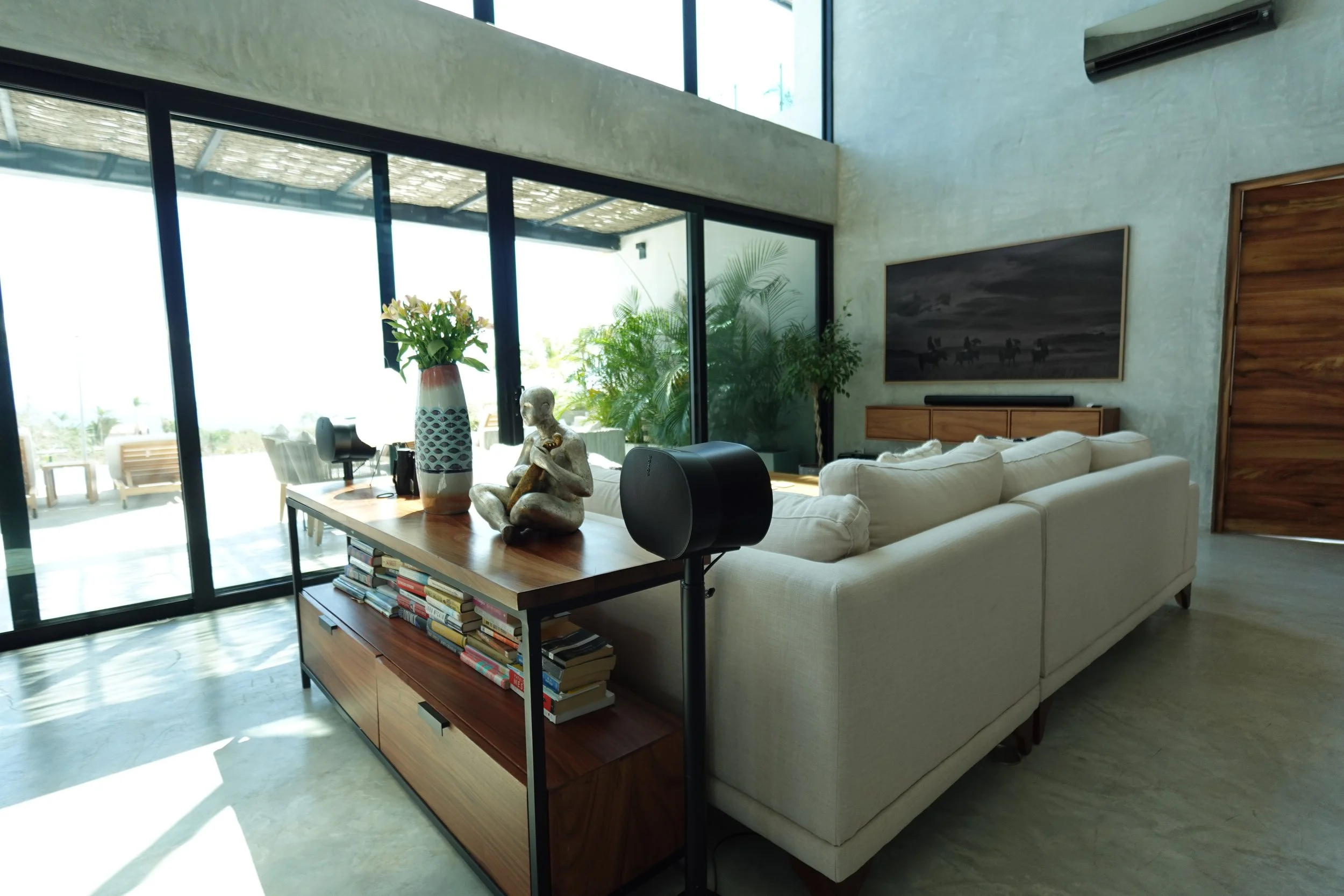 Living room with a beige sofa, a wooden coffee table with books, a tall vase with flowers, and decorative items including a statue and a speaker. Large floor-to-ceiling windows and a sliding glass door lead outside. There is a large framed black-and-