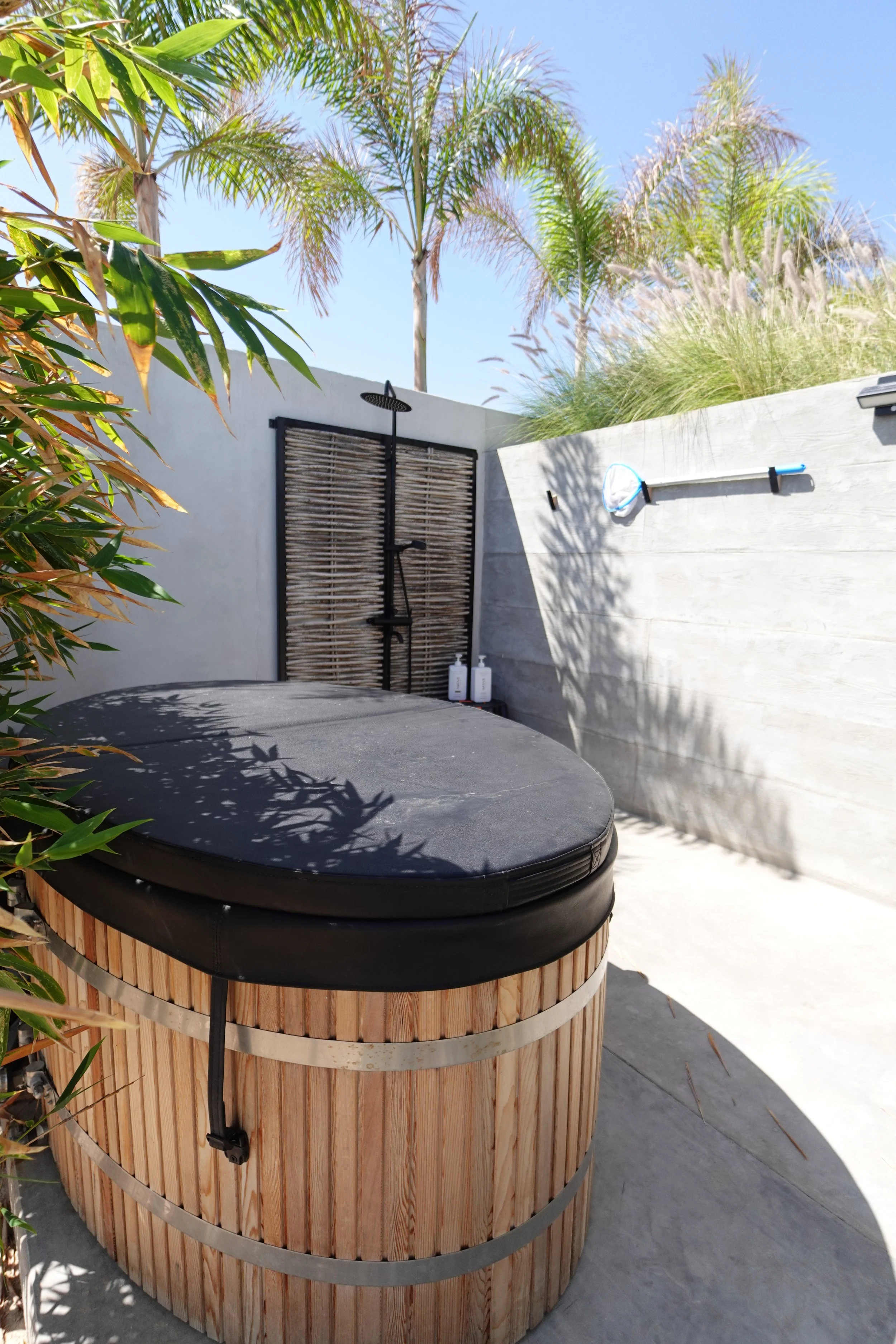 An outdoor shower area with a rain shower head, a bamboo privacy screen, and a wall-mounted tennis racket. There are several white bottles on the ground near the wall, with tall palm trees and grasses in the background, and a clear blue sky.