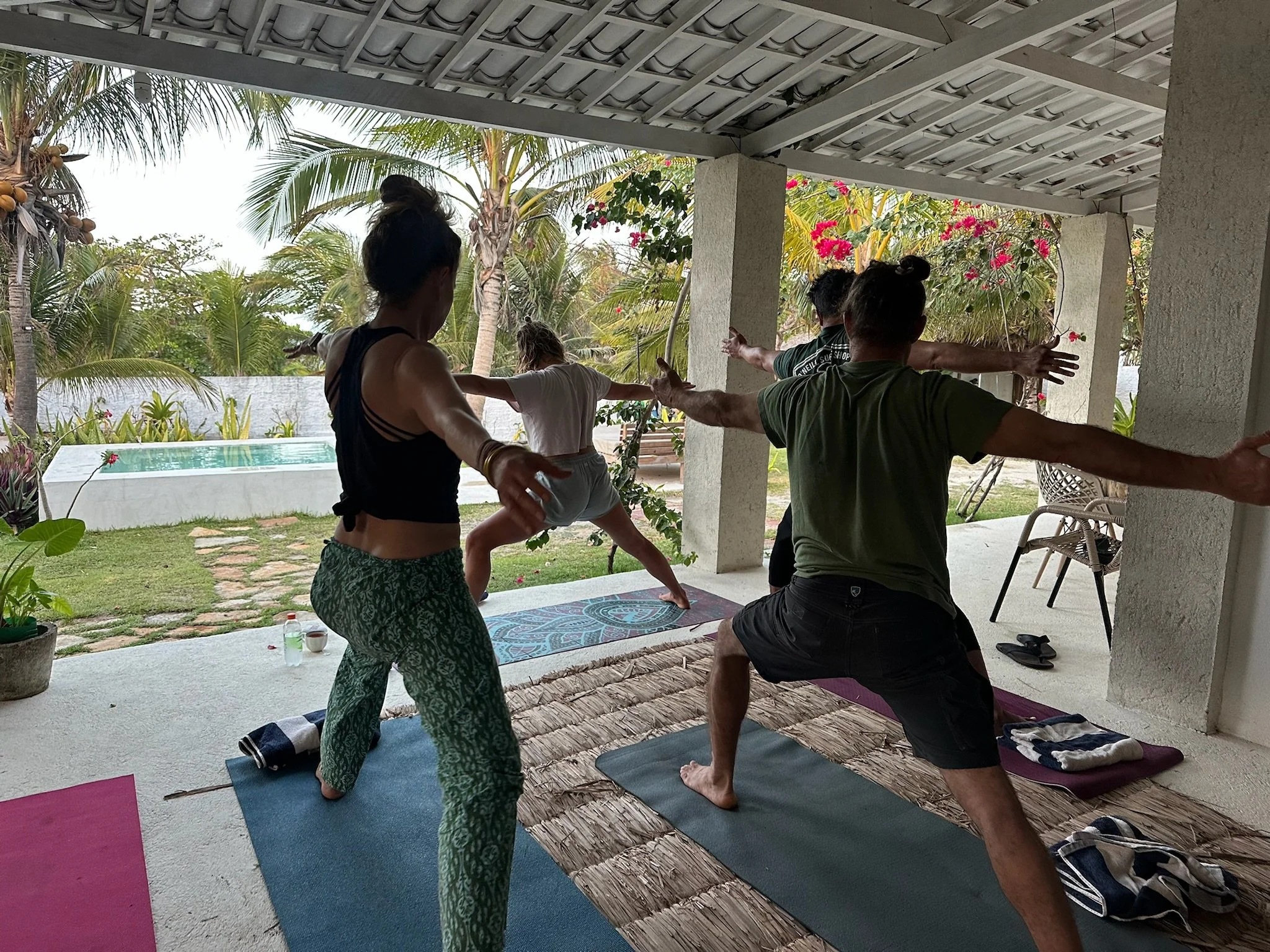 People practicing yoga on mats in an outdoor covered patio with tropical plants and palm trees in the background.