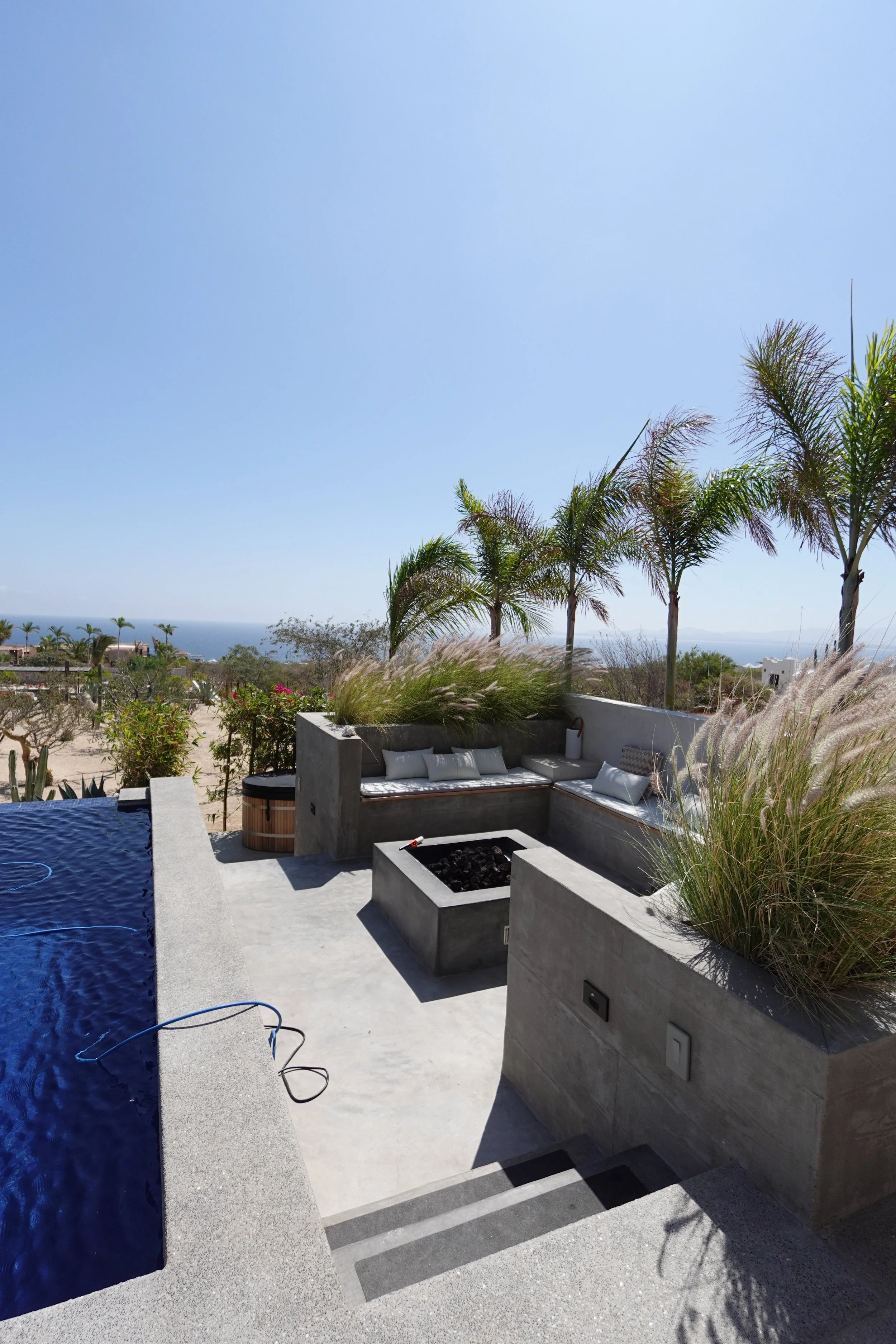 A luxurious outdoor patio with a swimming pool, palm trees, and a fireplace, overlooking a scenic view of the ocean and sky.