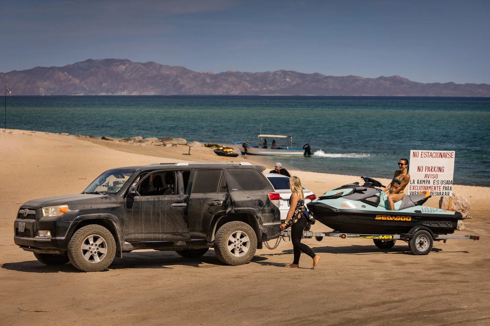 A beach scene with cars parked on the sand and people enjoying water activities near the shoreline. A woman in sunglasses and swimwear sits on a jet ski, and others are near a boat in the water. Mountains are visible in the background.
