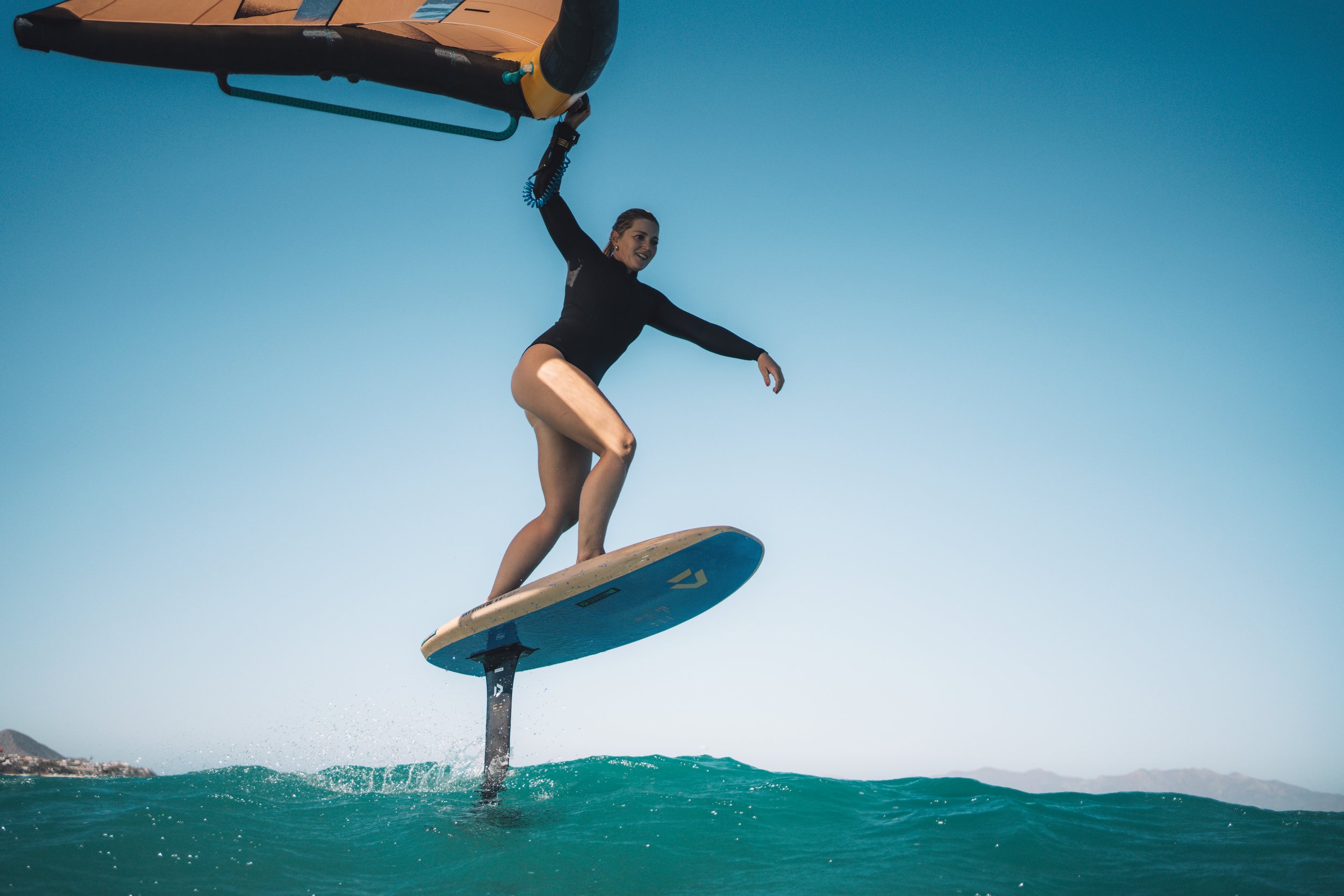 A woman on a hydrofoil surfing on the ocean waves during clear weather, with a distant shoreline and mountains in the background.