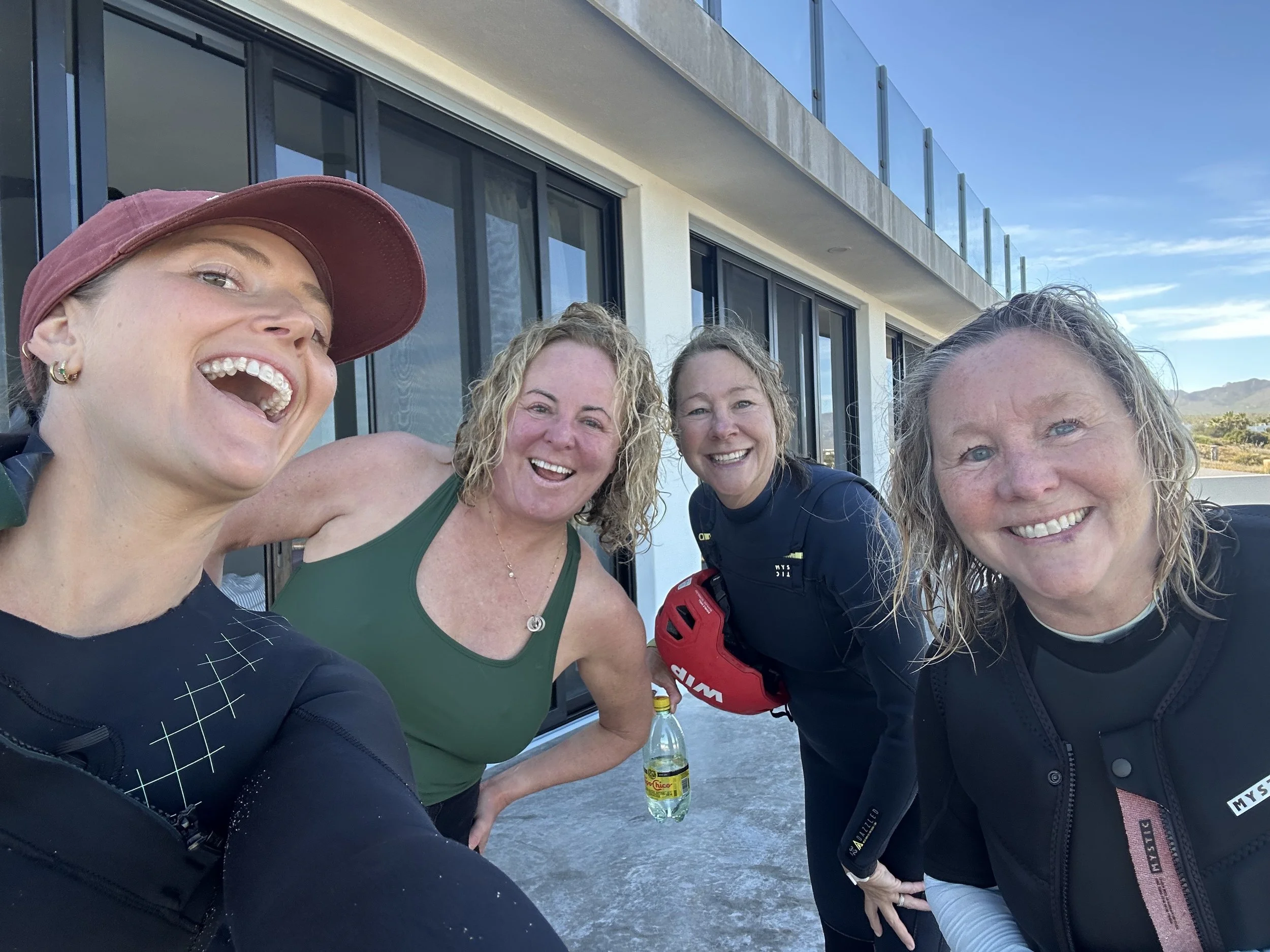 Four women smiling and posing closely together outside in front of a modern building with large glass doors and windows, with a cloudy blue sky and distant mountains in the background.