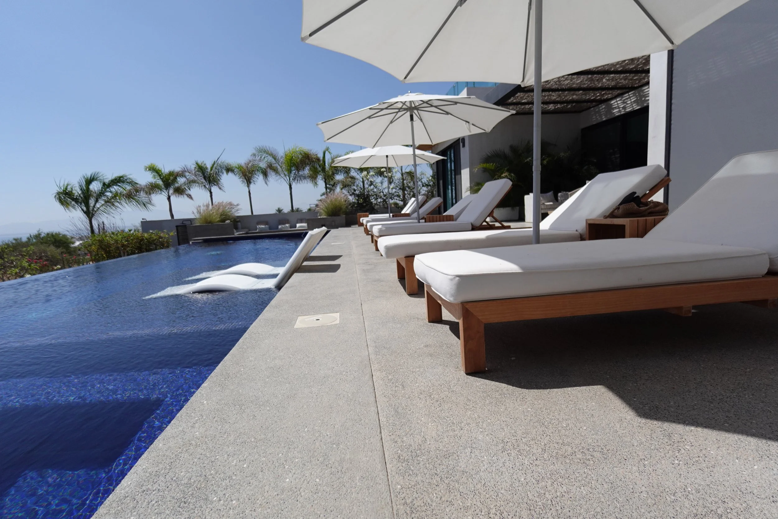 A luxurious outdoor pool area with pool chairs, umbrellas, and a view of palm trees under a clear blue sky.