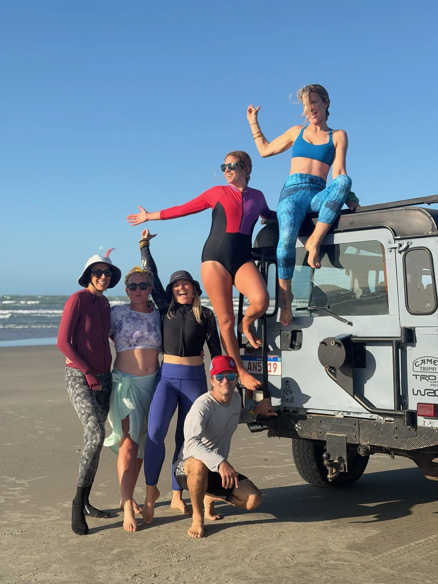A group of people on a beach with a vehicle, some wearing athletic and beachwear, enjoying a sunny day.