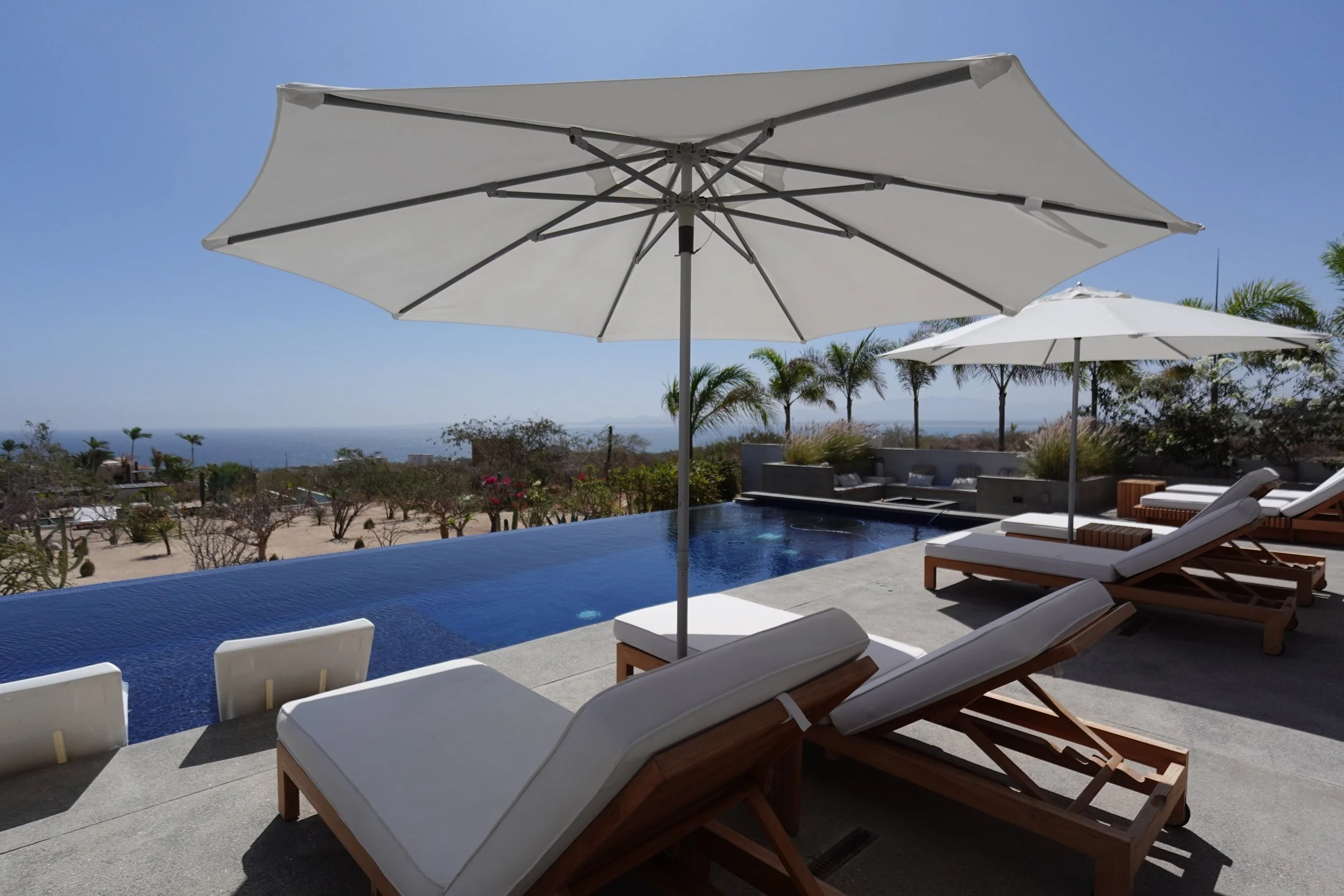 Outdoor poolside area with white lounge chairs, large white umbrellas, and a view of a distant ocean or sea under a clear sky.